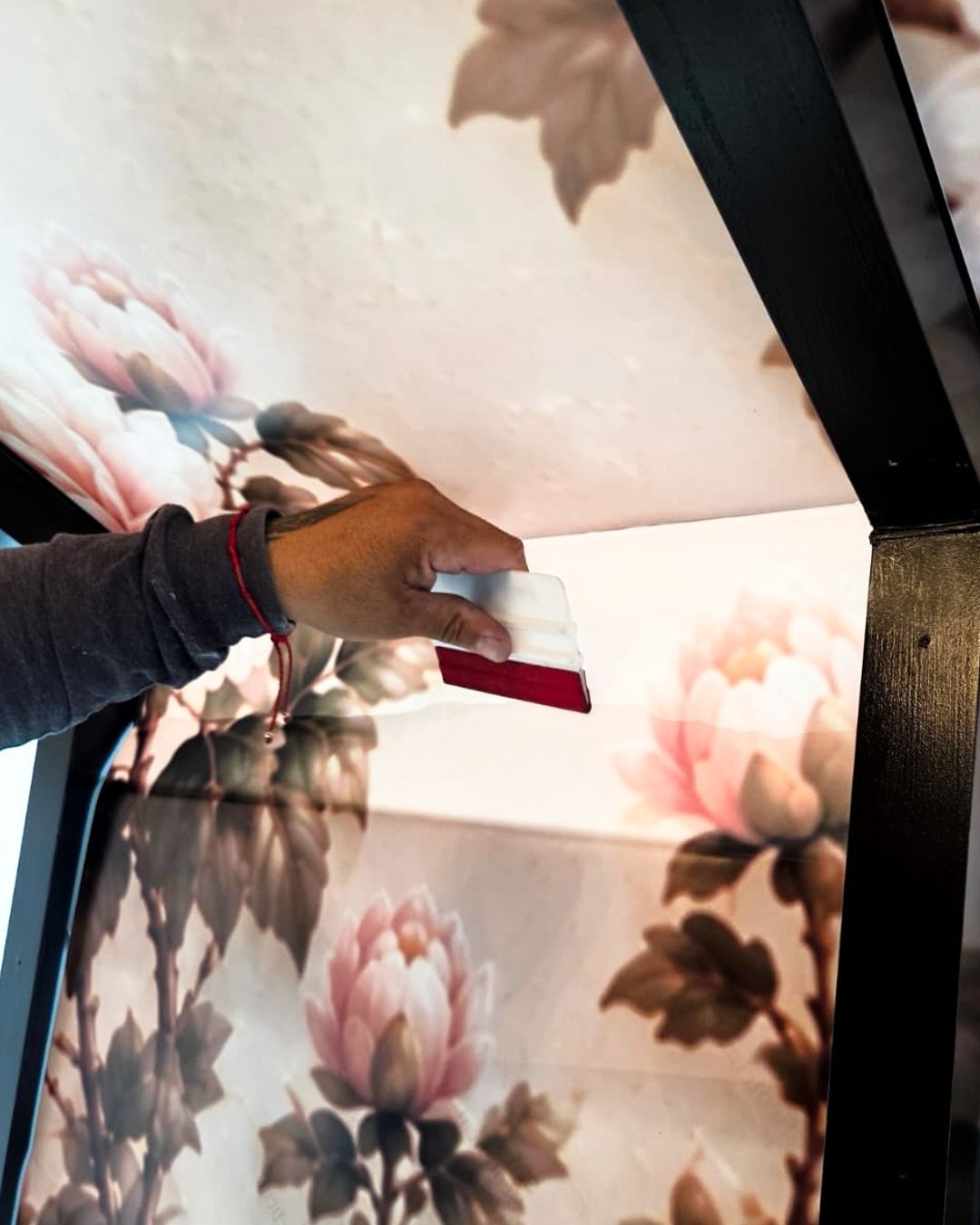 Hand holding a white-and-red spray bottle near floral wallpaper and a black metal frame.