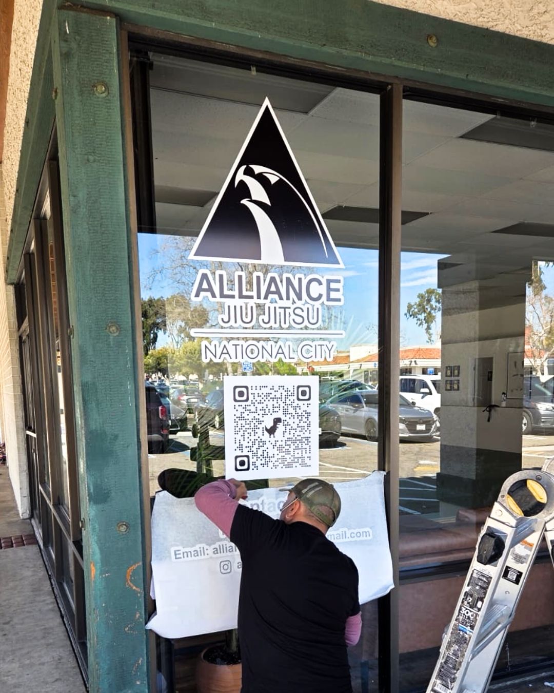 Person applying a sign to a glass storefront door with the Alliance Jiu Jitsu logo and QR code.