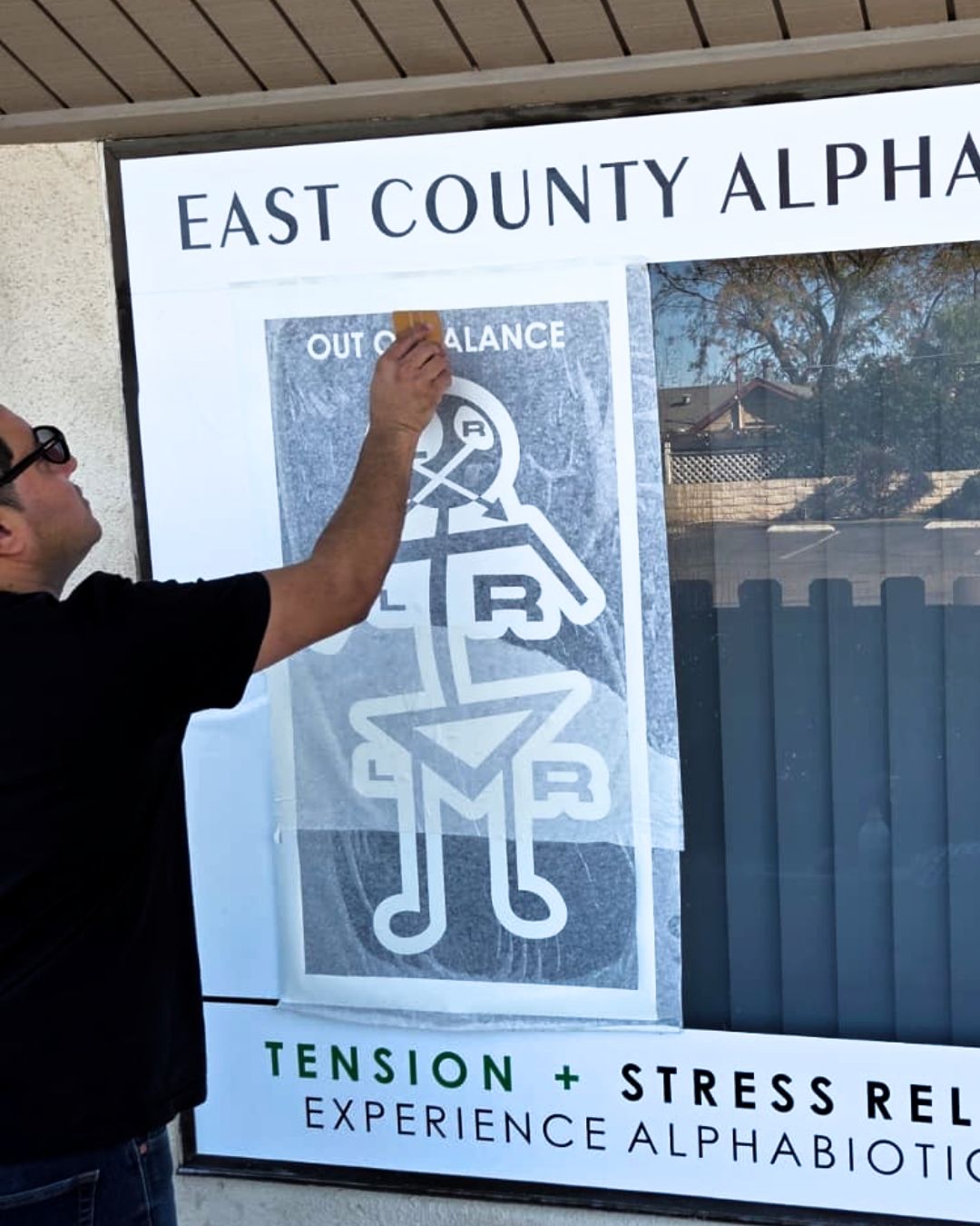 A person applying a poster to a storefront window, reading East County Alpha and Tension + Stress Relief.