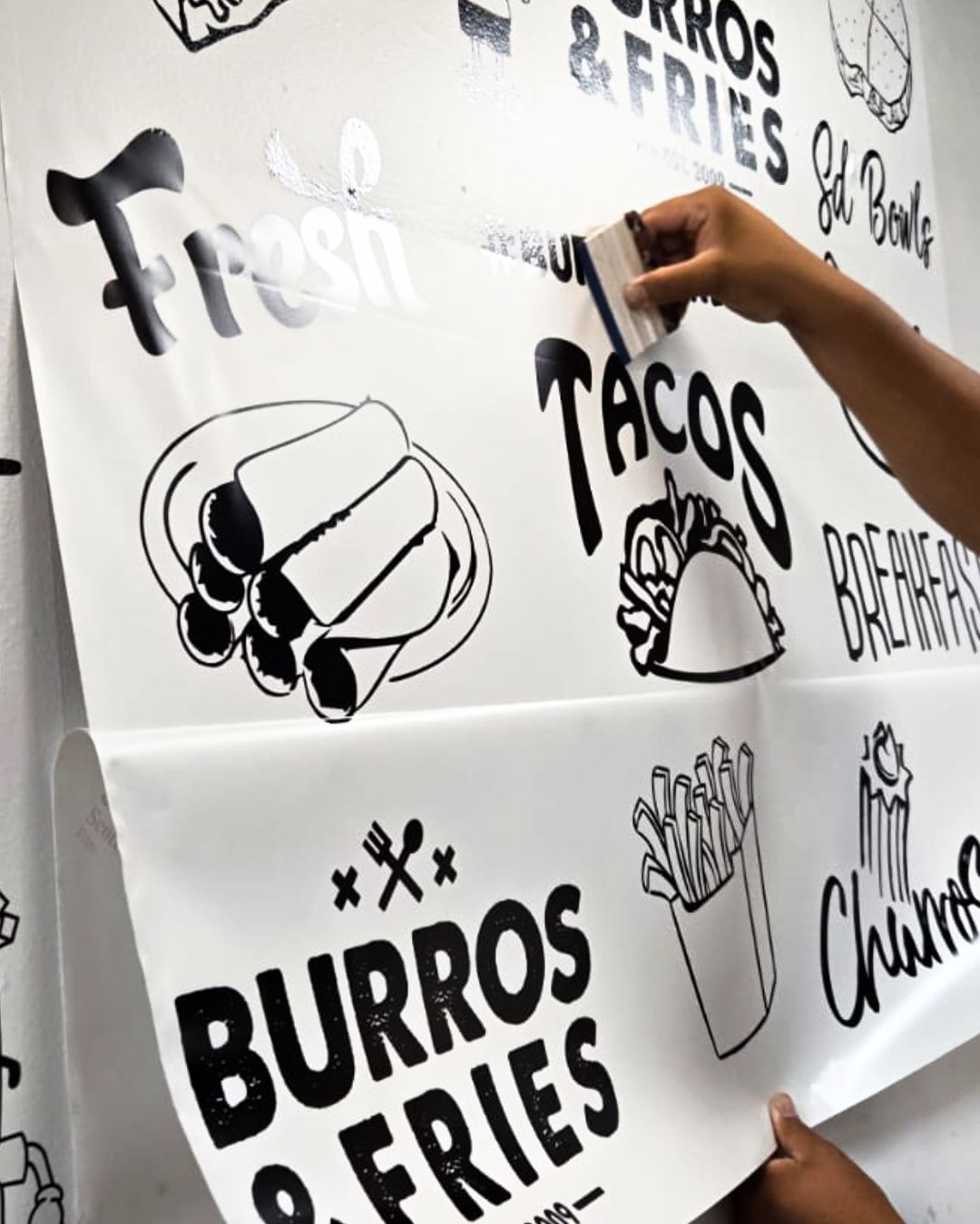 Person painting a white food menu sign with bold black lettering for tacos, burritos, and fries.