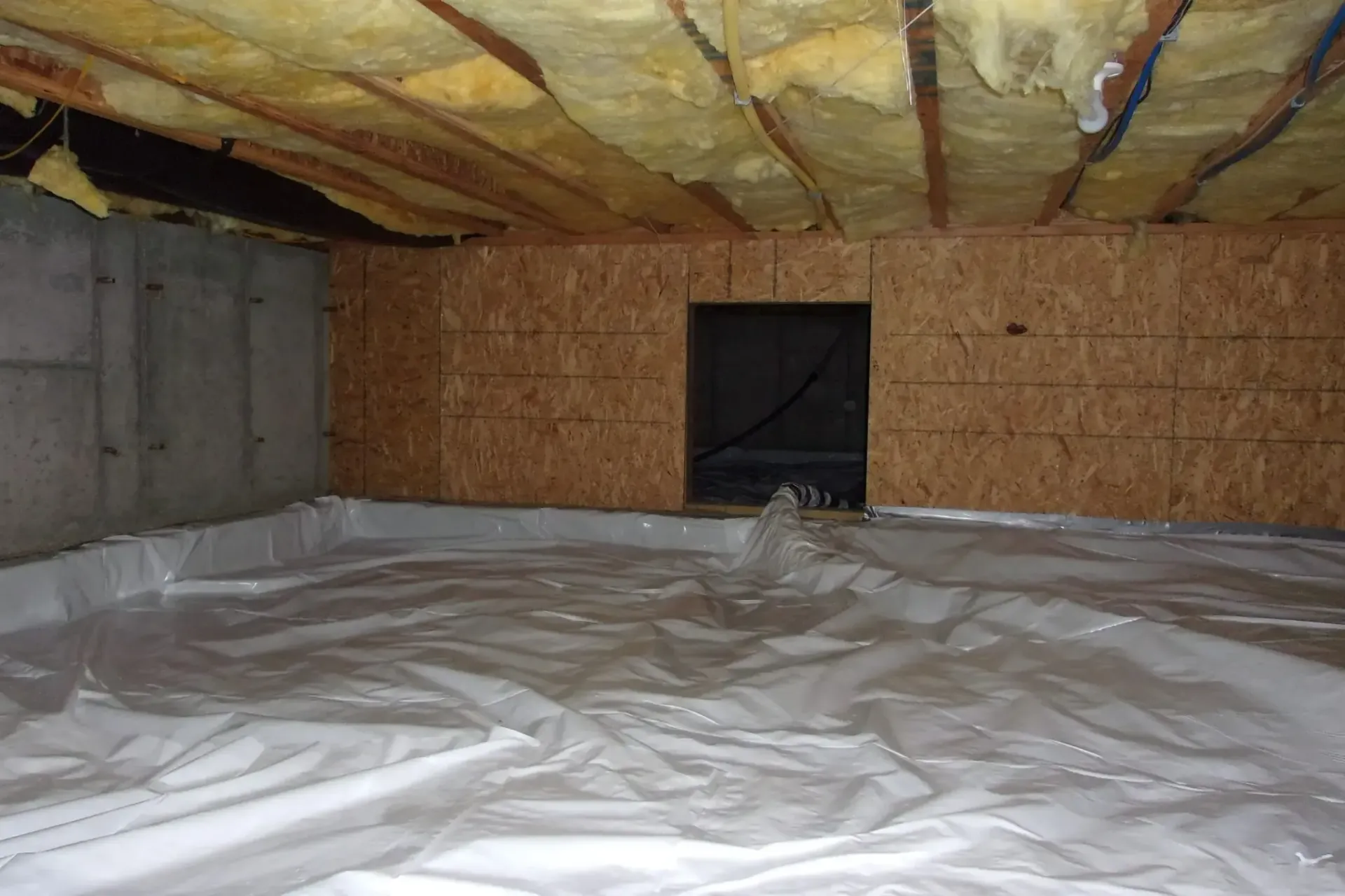 Unfinished basement with plastic-covered floor, exposed insulation ceiling, and a small window in the back wall