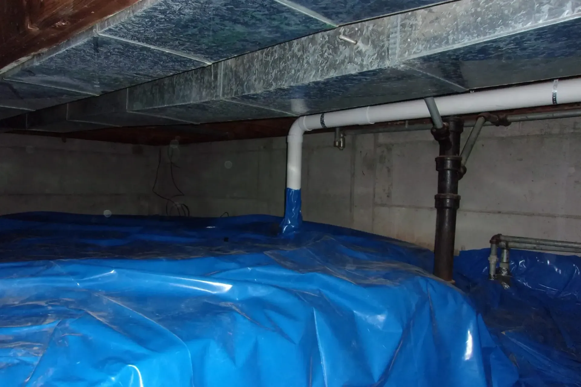 Blue tarp covering items in a low crawlspace under a house, with exposed pipes and beams.
