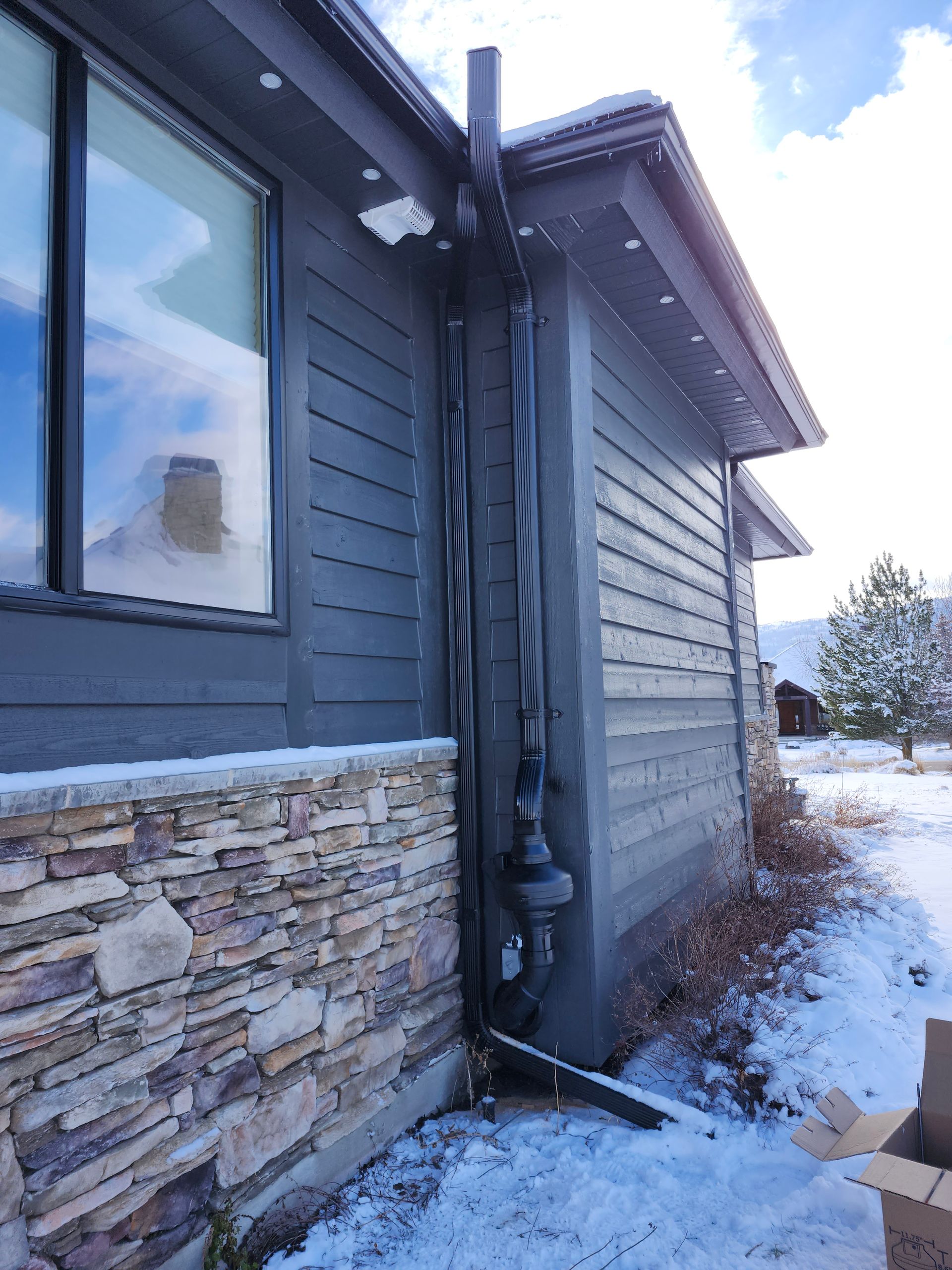 Snowy exterior corner of a house with gray siding, stone trim, and downspouts