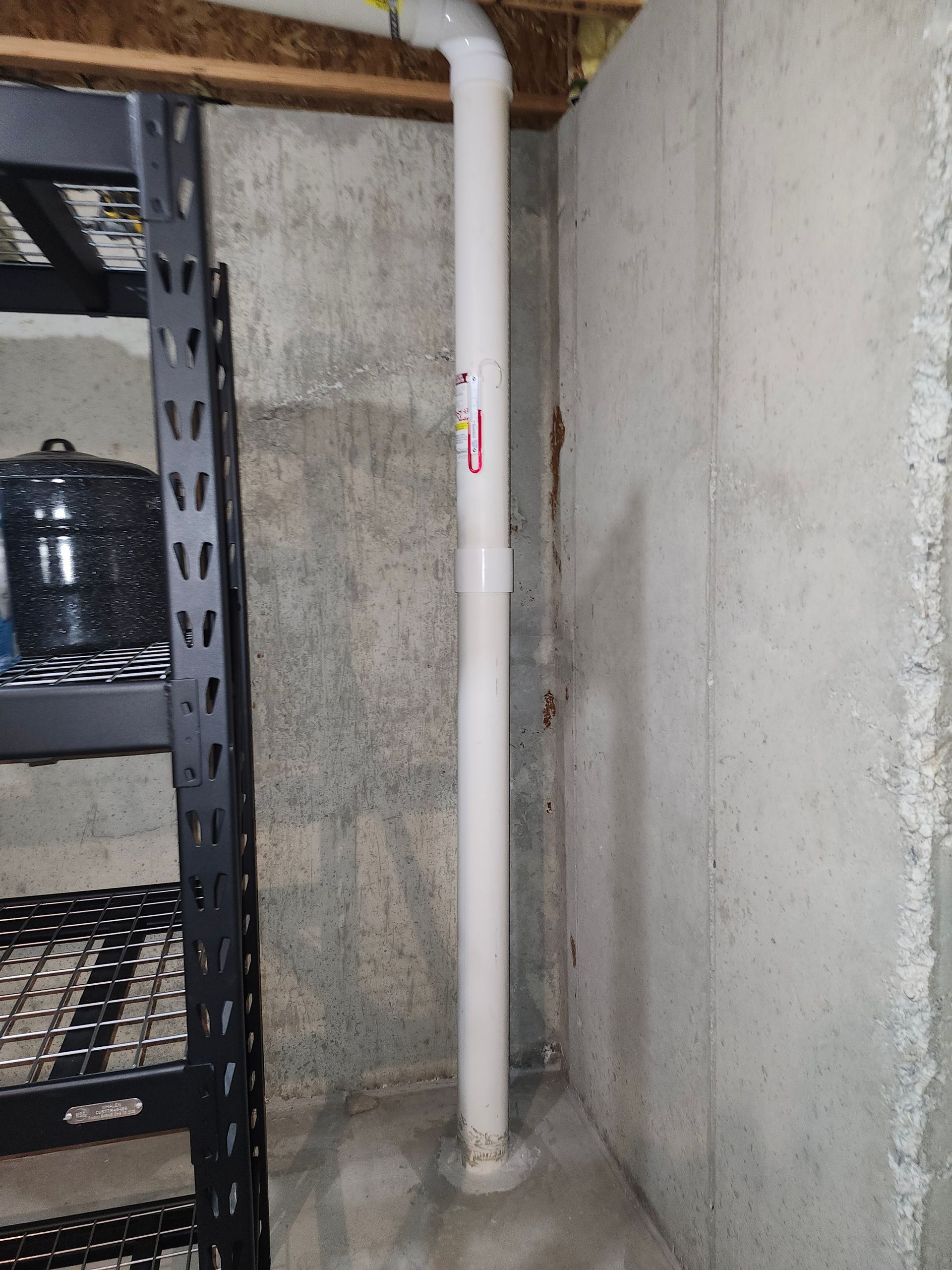 Basement utility corner with a white PVC pipe beside a black metal shelving unit.