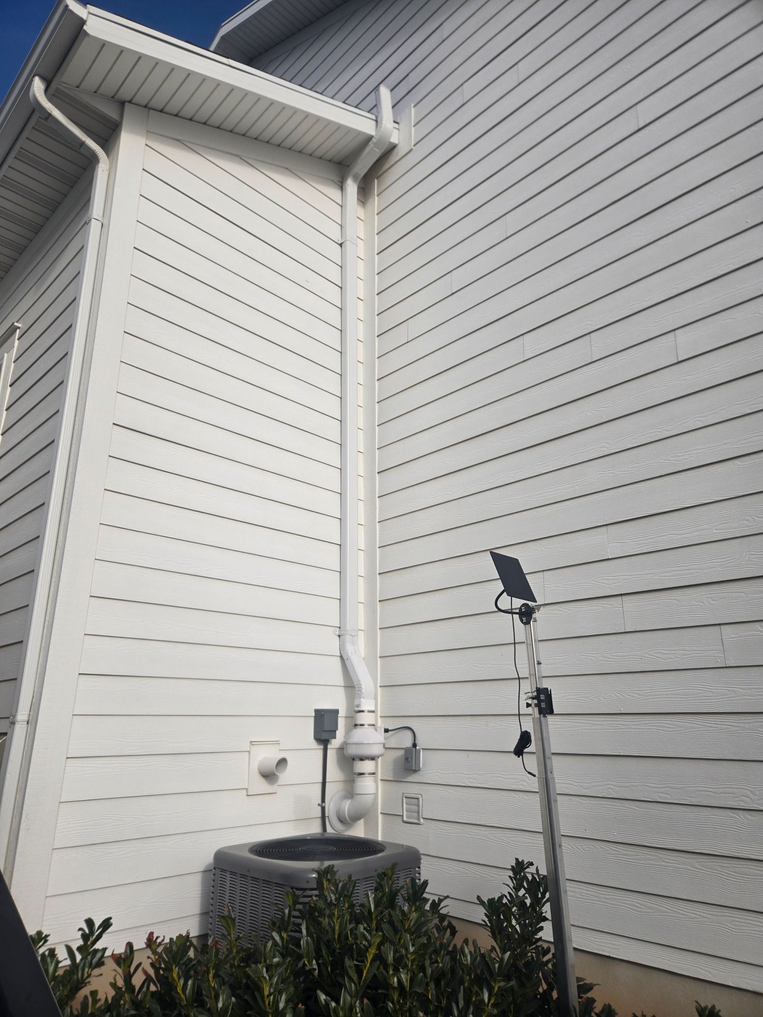 Outdoor air conditioner unit beside a white house wall and downspout under an overhang
