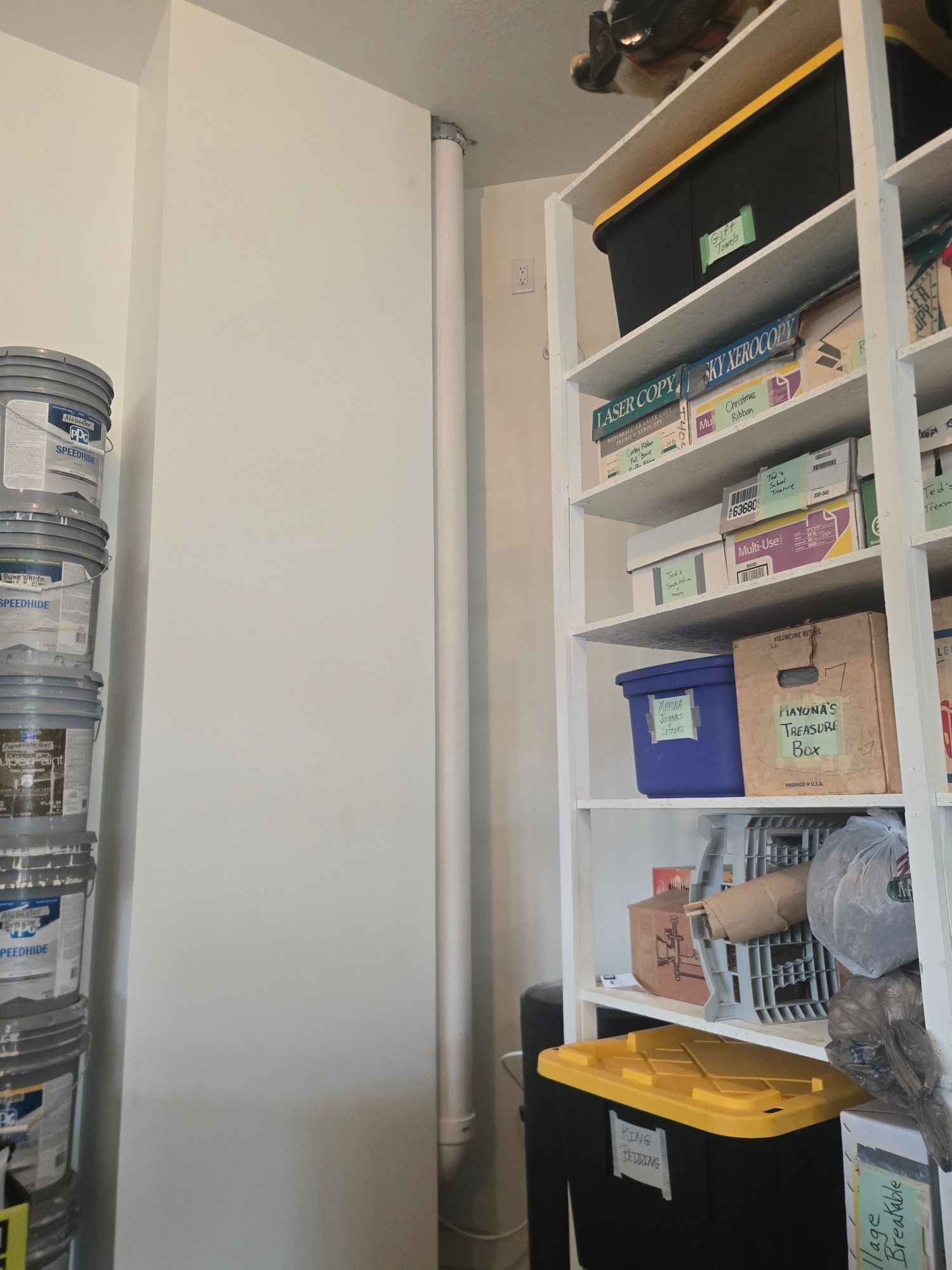 Closet shelf with stacked storage bins, supplies, and a yellow-lidded bin beside a white cabinet