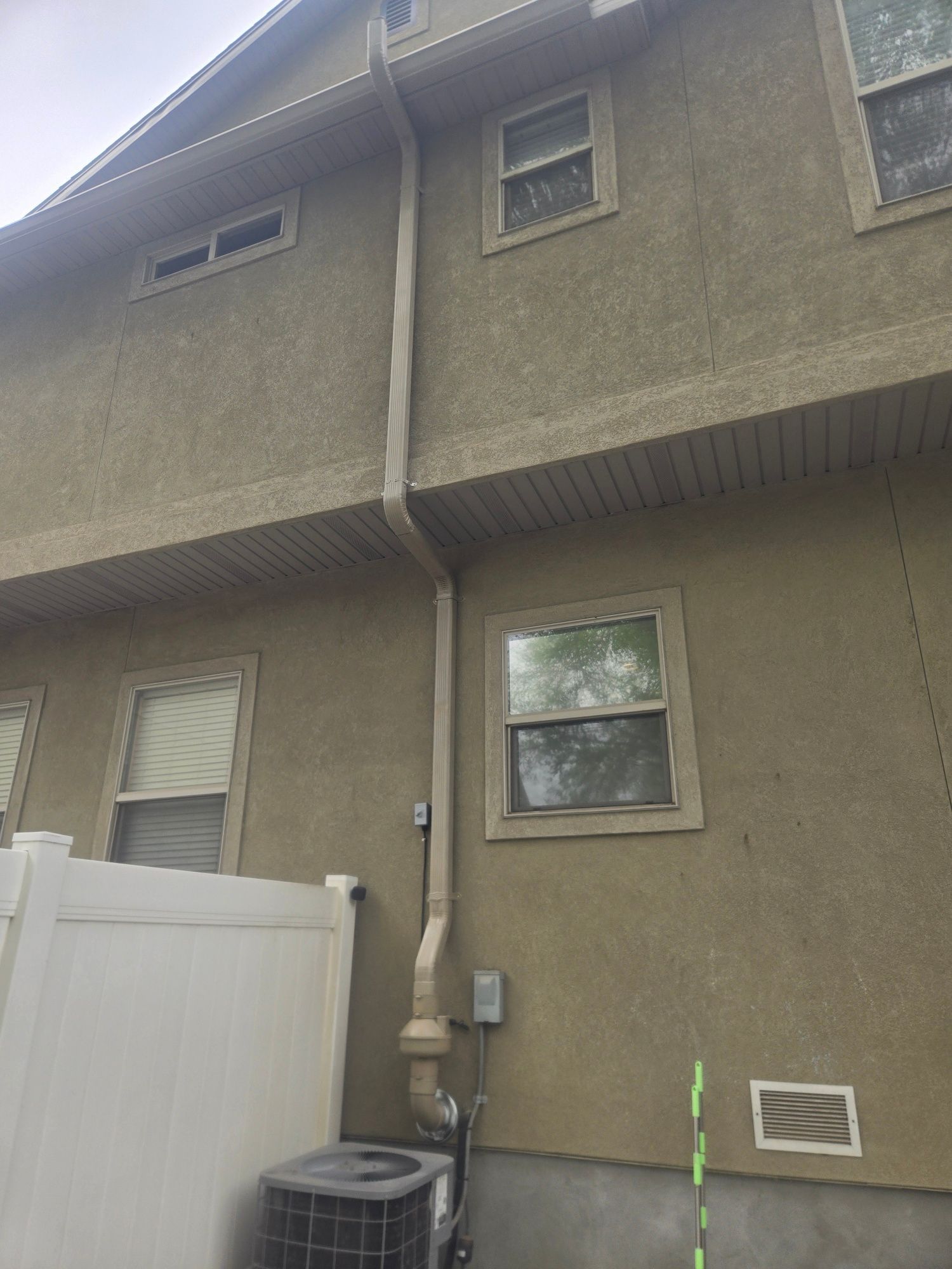 Exterior wall of a two-story house with windows, drainpipe, and white fence below.