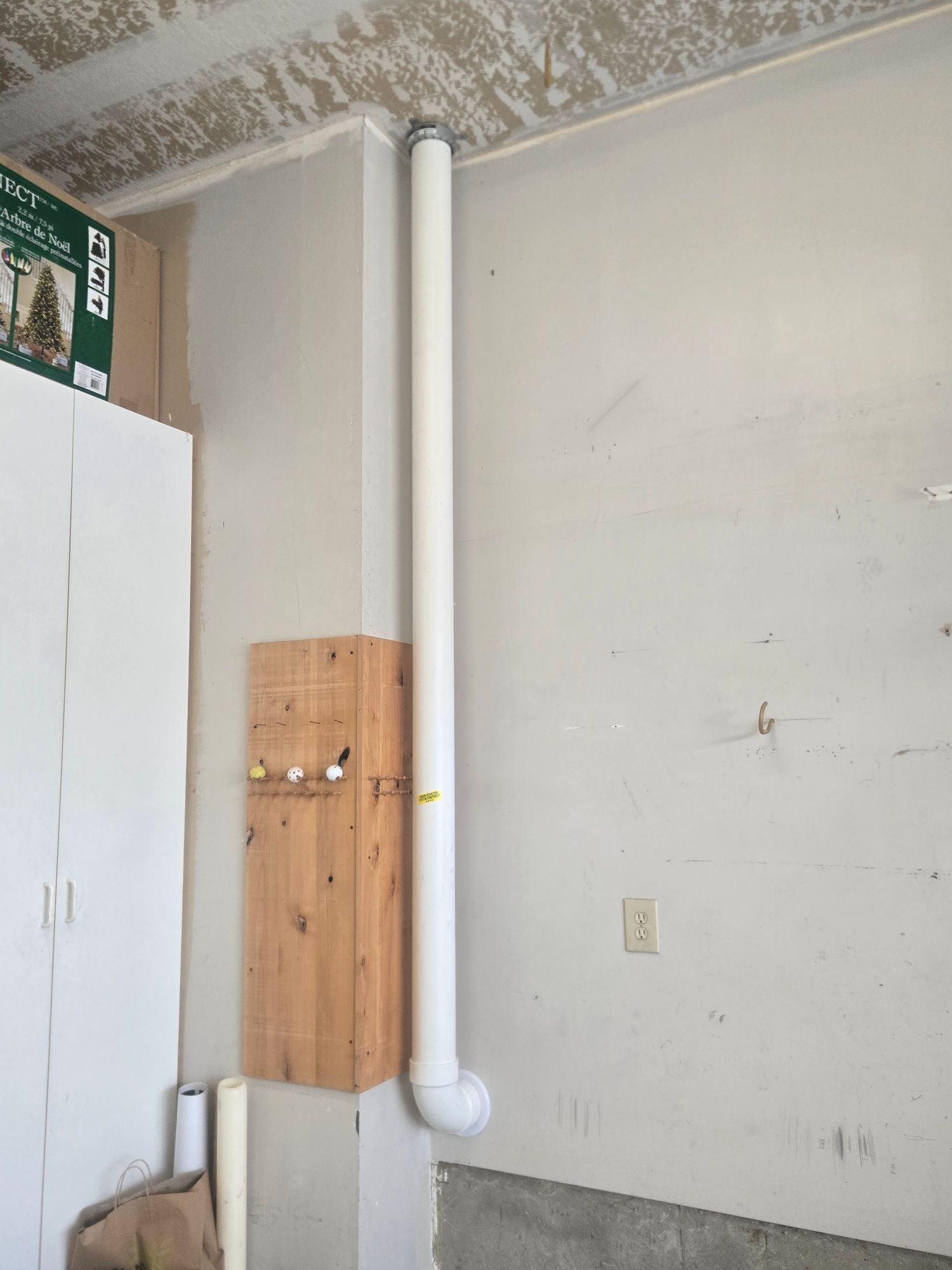 Utility room corner with white cabinet, wall vent pipe, and wooden board mounted on a beige wall.