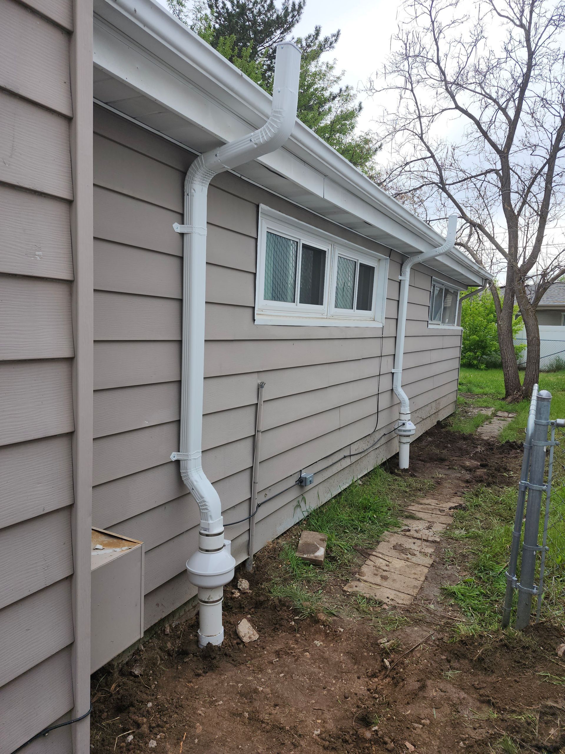 Side of beige house with white gutters and small windows, next to a dirt strip and chain-link fence.