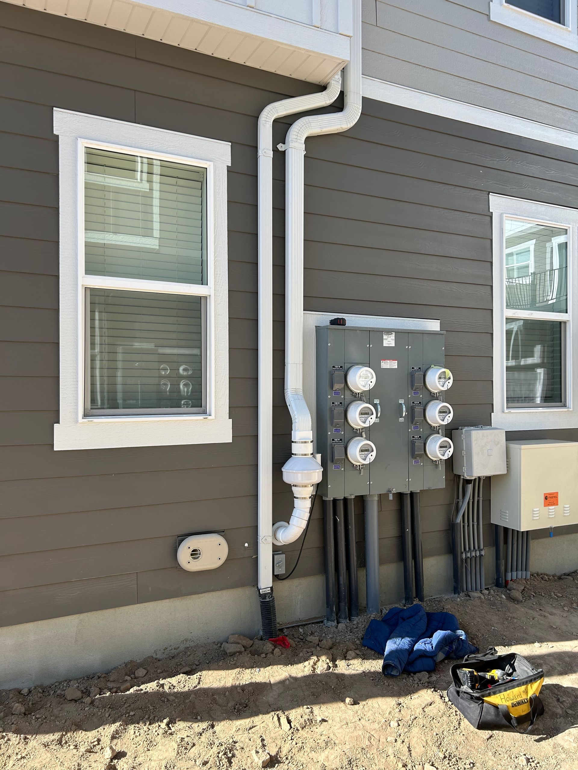 Exterior wall with utility meters and pipes beside two windows, gray siding, and a small dirt area.