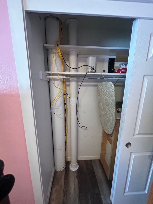 Closet utility area with white pipes, shelving, and a standing ironing board beside an open door.