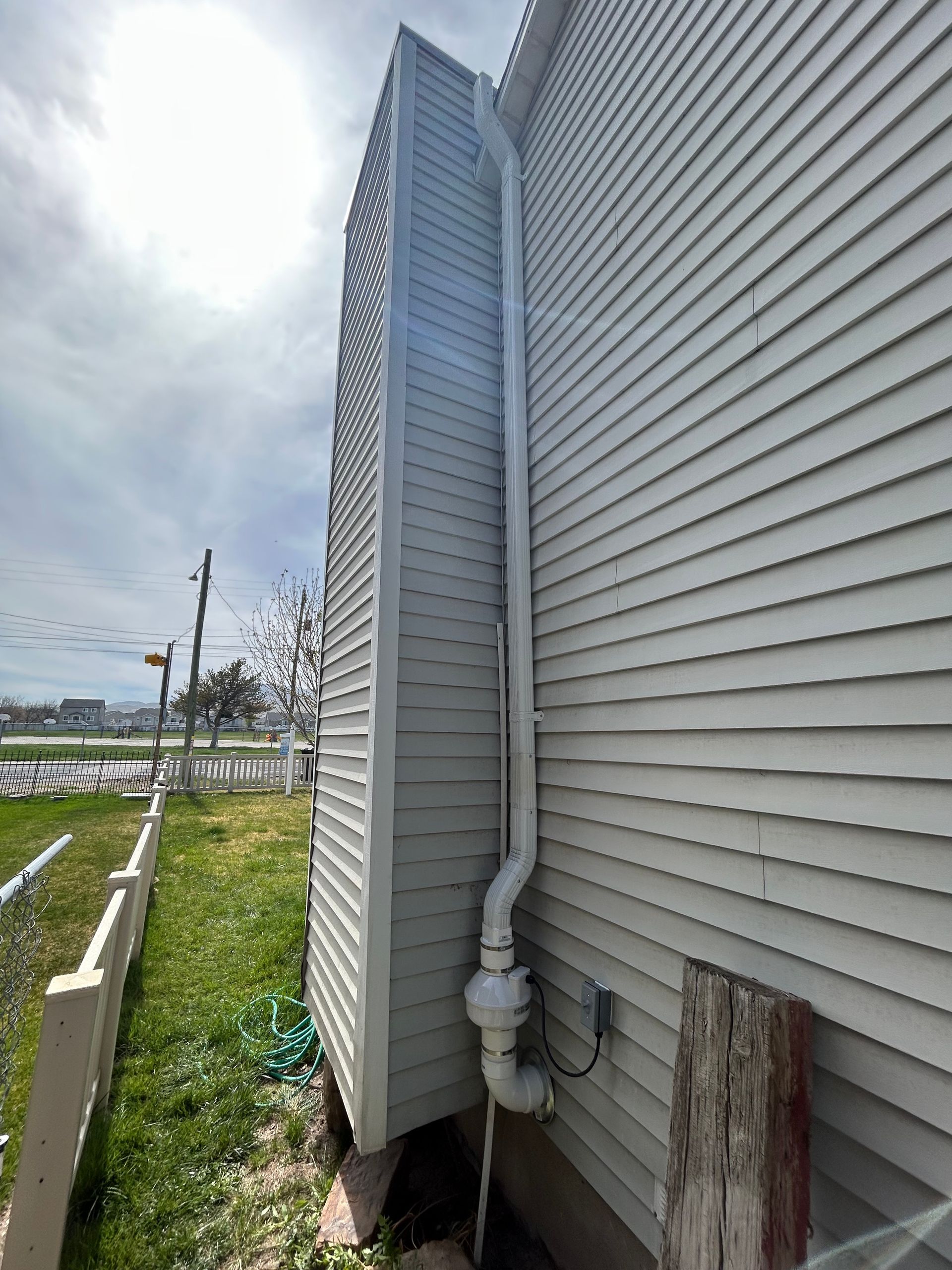 White-sided building corner with gray vent pipe, beside a grassy roadside under a cloudy sky