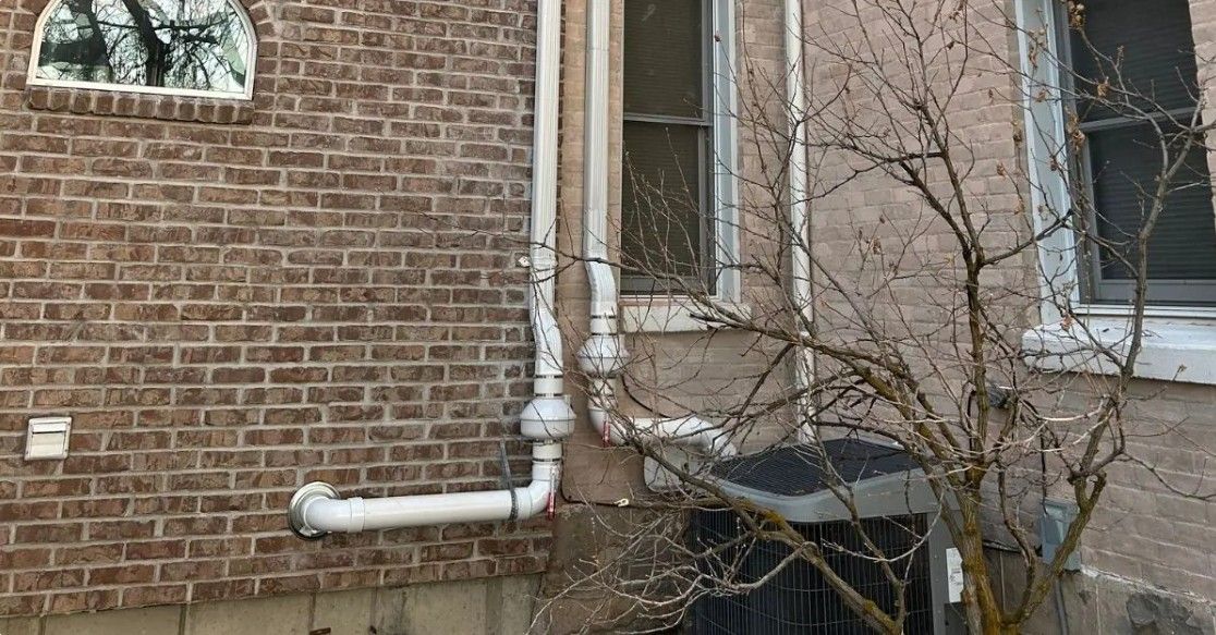 Brick house exterior with white pipes, a window, and a leafless tree by the wall