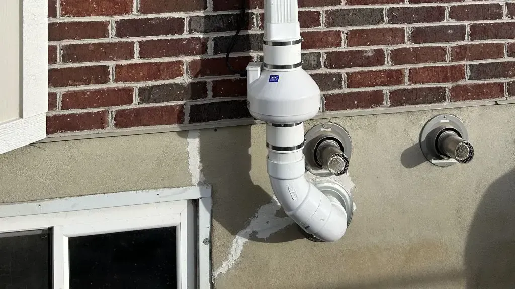 White plumbing vent pipe and two wall outlets on a brick-and-stucco exterior wall above a basement window
