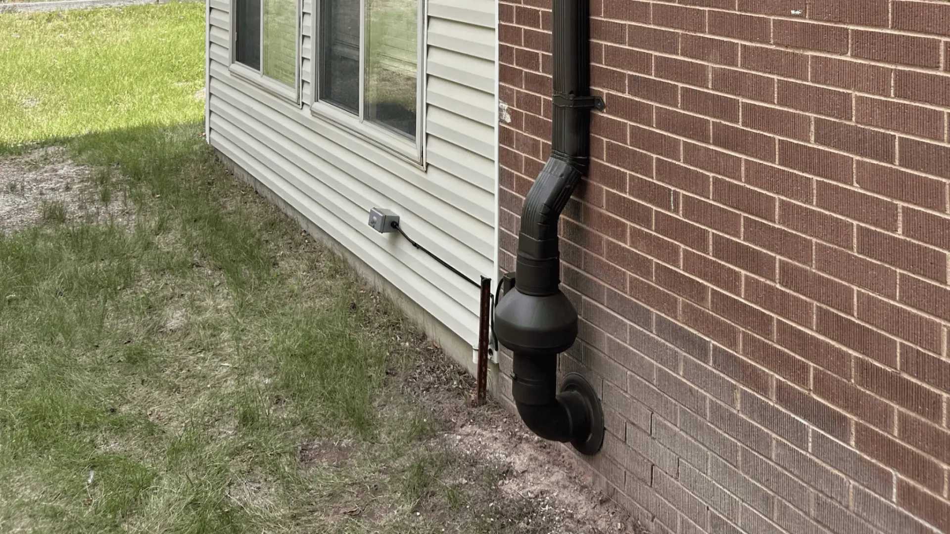 Black downspout and pipe beside a brick house with beige siding and grassy yard.