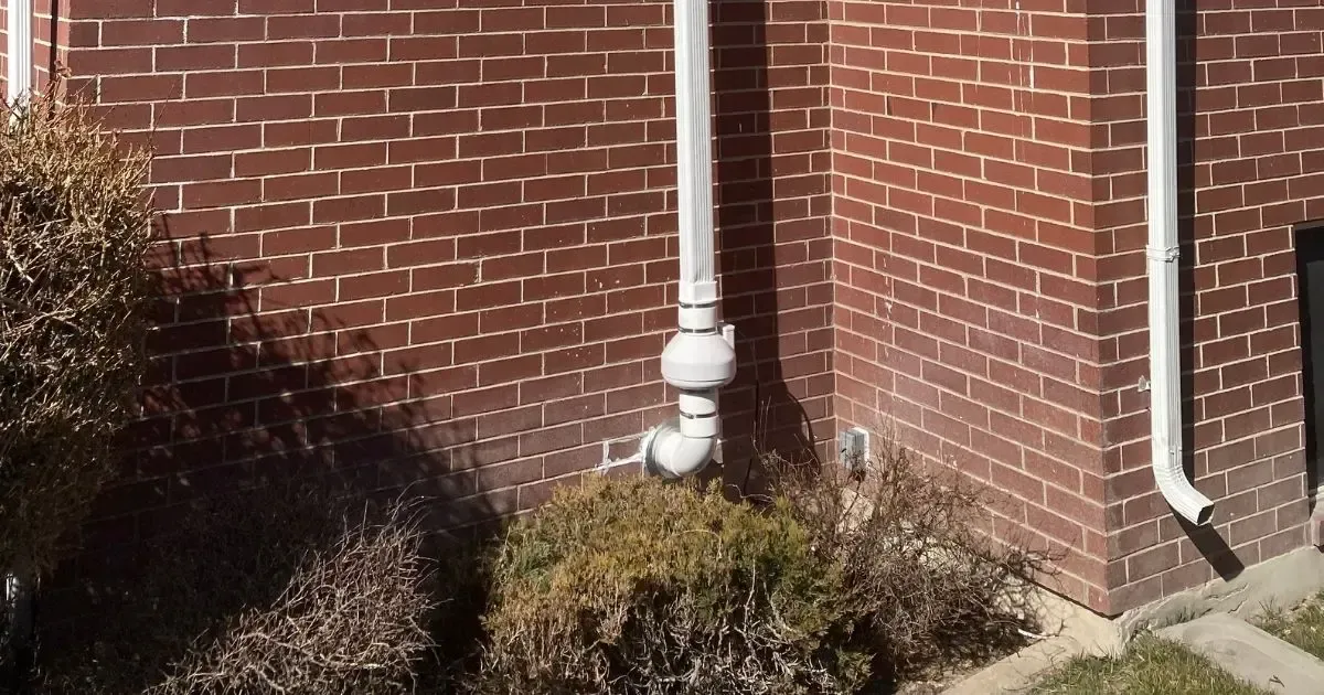 White gutter downspout on a red brick house beside shrubs and a narrow concrete walkway.