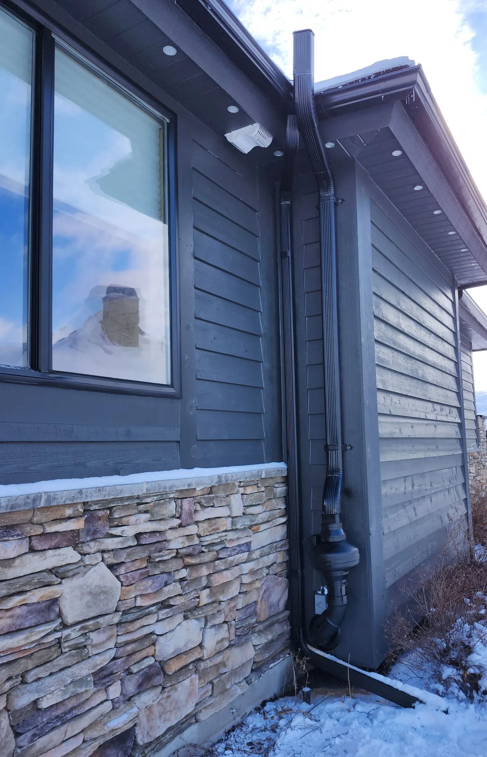 Snow-covered side view of a gray house with stone siding, windows, and downspouts in winter.