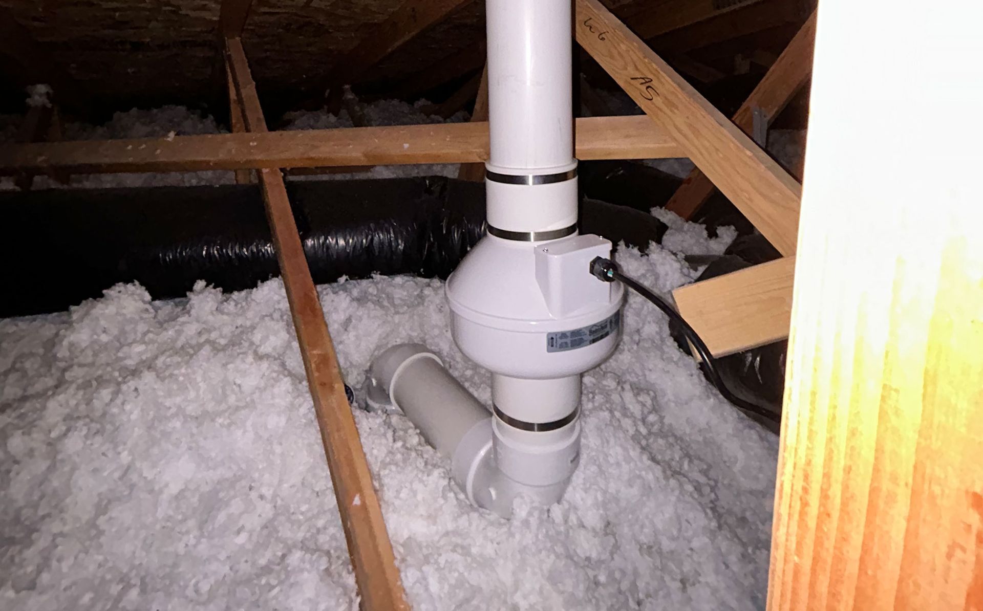 Attic with white PVC vent pipe extending through loose insulation and wooden rafters