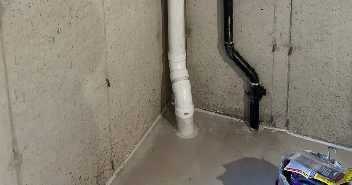 Concrete wall corner with white and black pipes and a small wet floor puddle.