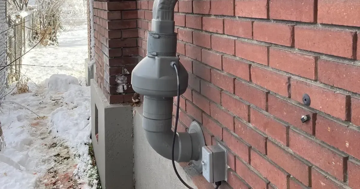 Gray vent pipe and utility box mounted on a red brick wall beside a snowy outdoor path.