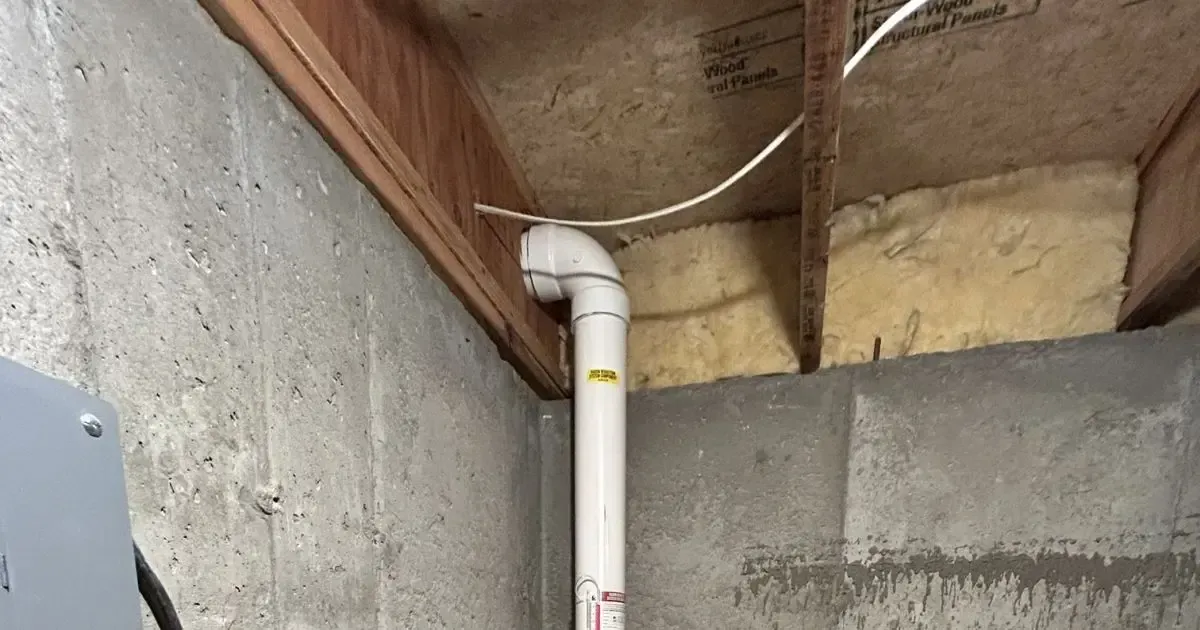Basement corner with white PVC pipe, concrete walls, exposed insulation, and wood framing.