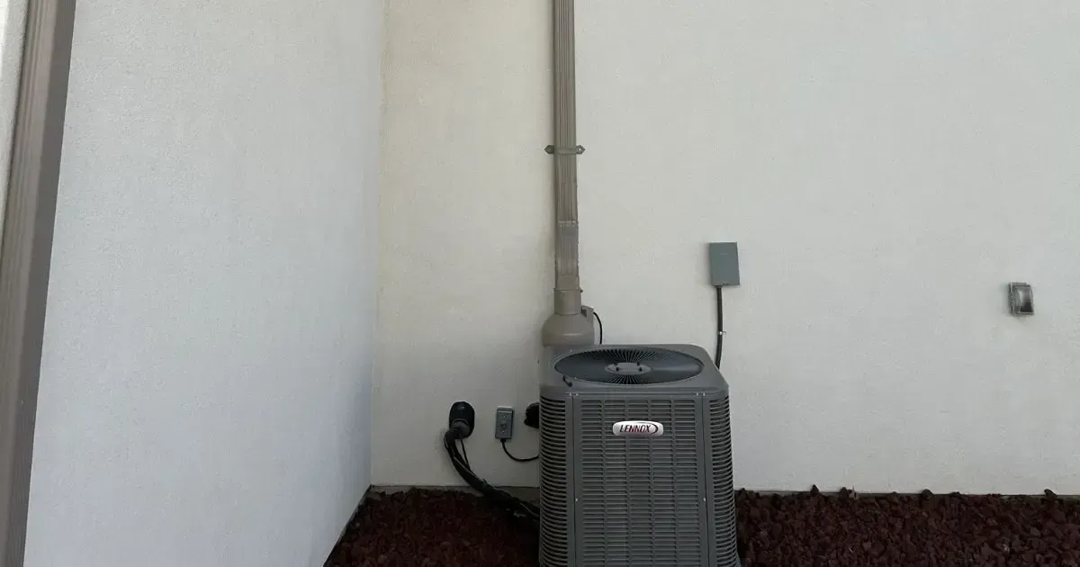Outdoor HVAC unit beside a white wall with a vertical pipe and electrical box above it
