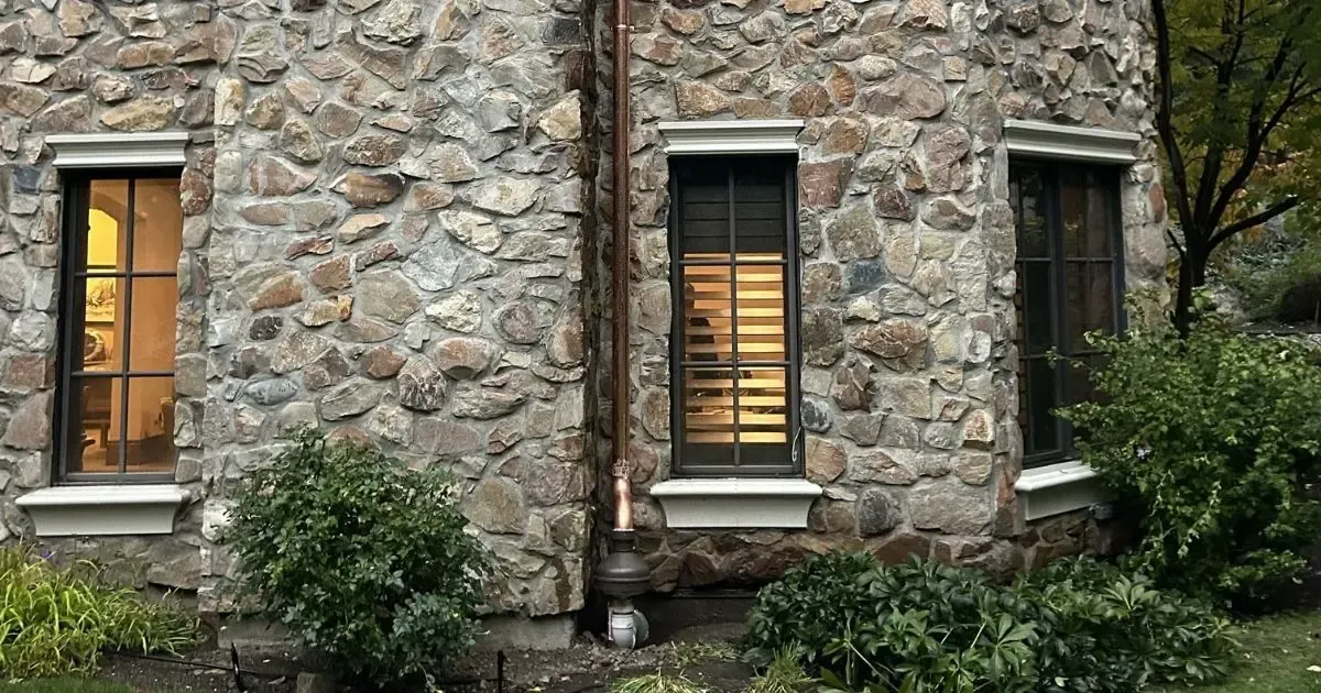 Stone house wall with three lit windows and surrounding shrubs