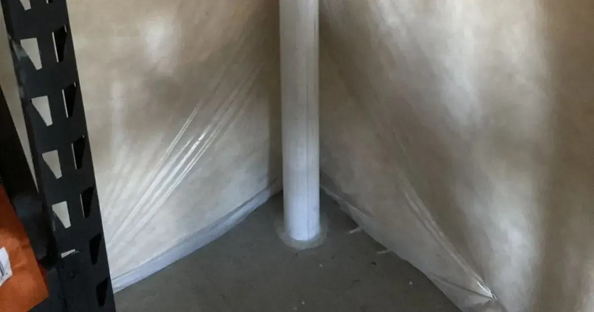 Corner of a room with a gray floor, beige walls, and a white pipe near metal shelving.