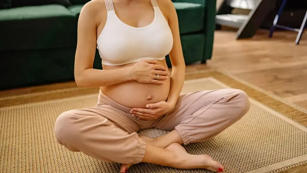 Pregnant person sitting cross-legged on a rug, holding their belly in a cozy living room.