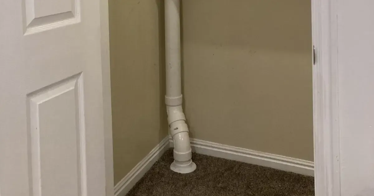 White PVC drain pipe in a beige hallway corner beside a white door and carpeted floor.