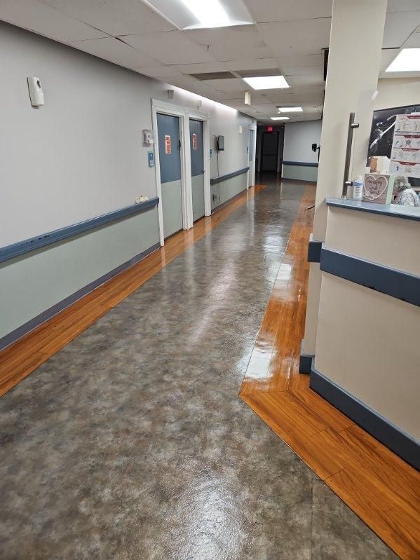Hallway in a medical facility with gray and wood-toned flooring, white and gray walls, and several closed doors.