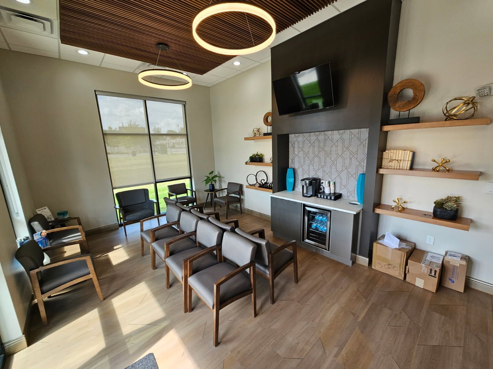 Waiting room with rows of chairs, coffee station, and a TV mounted on a dark wall with decorative shelves.
