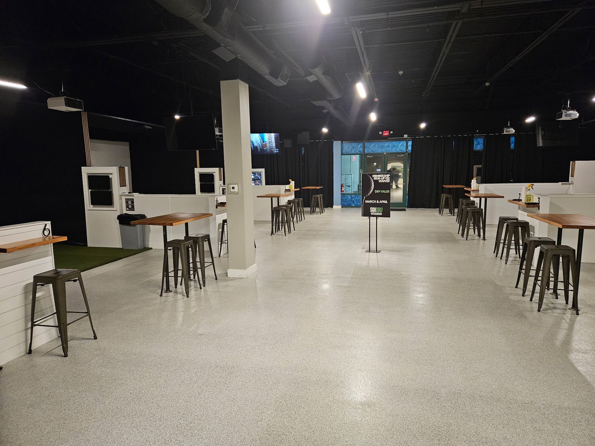 Indoor event space with tables, stools, and screens, dark walls, and light speckled floor.