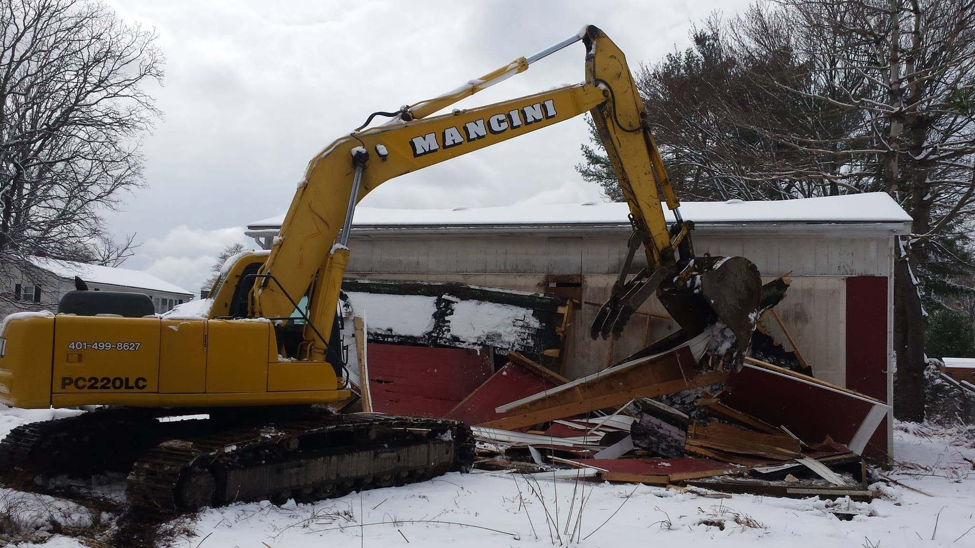 Yellow excavator demolishing a red garage in snowy yard