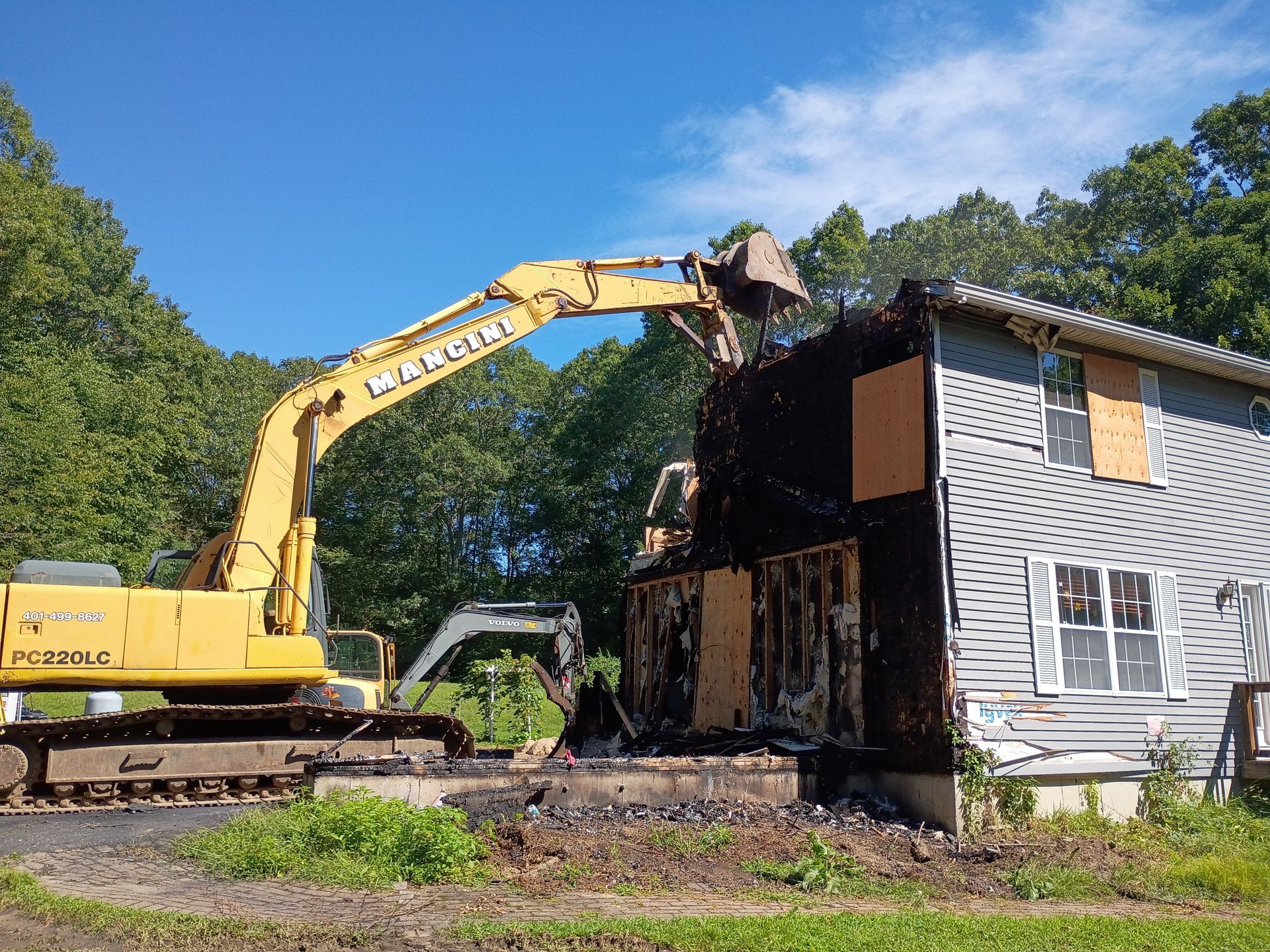Yellow excavator demolishing a burned two-story house under a clear blue sky