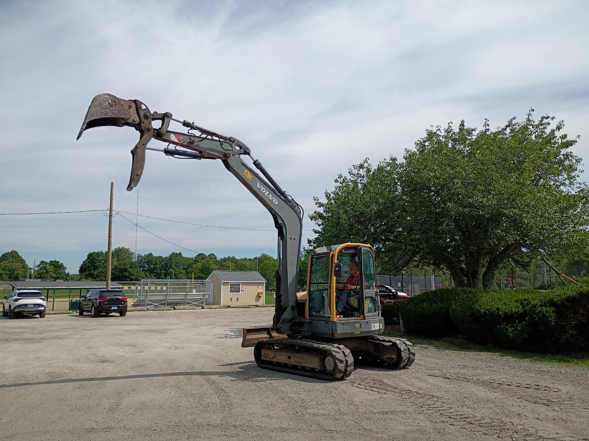 Excavator with raised claw on a gravel lot under a cloudy sky, trees and parked cars behind it