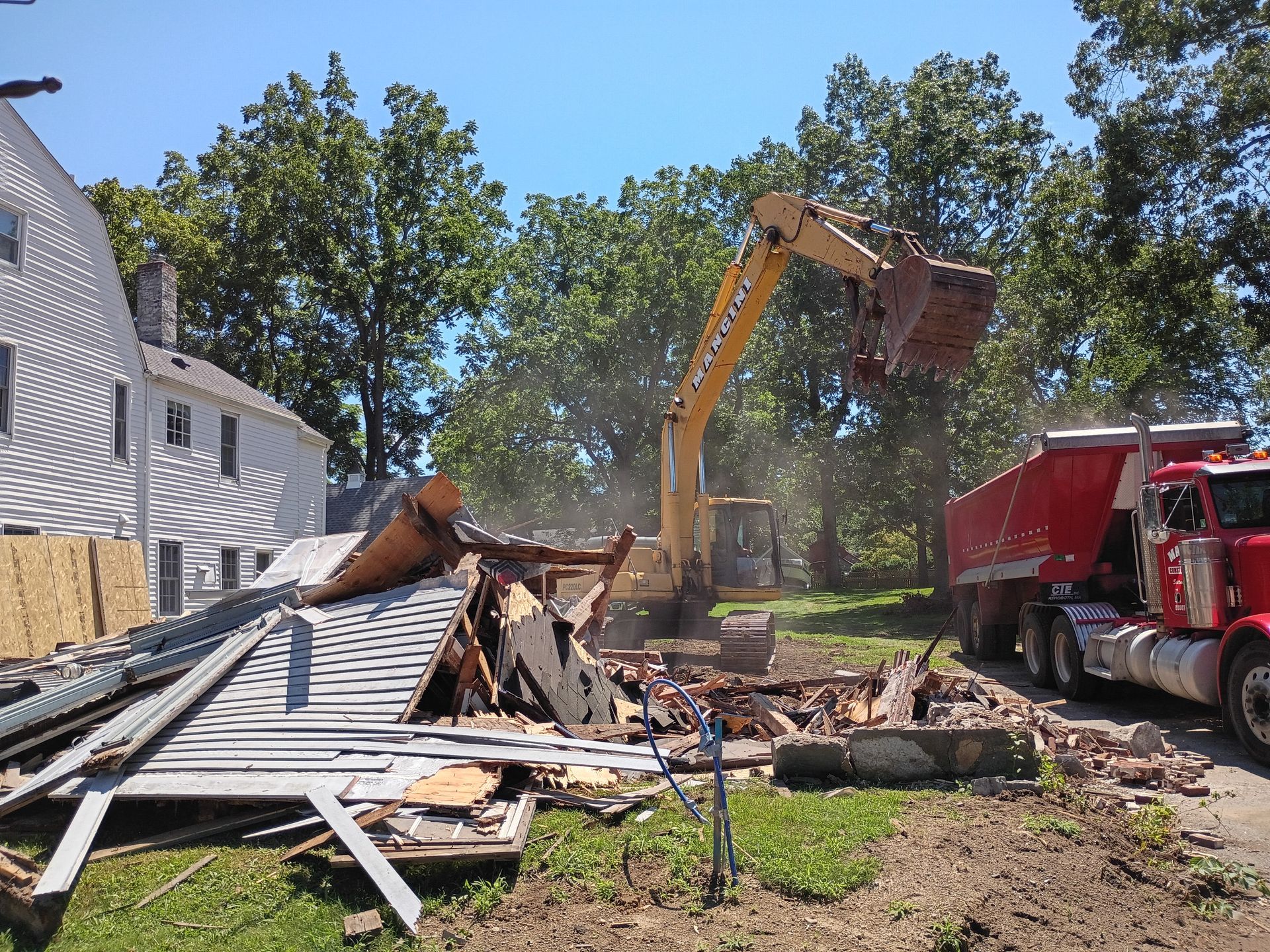 Excavator and dump truck clearing debris from a collapsed building beside a white house