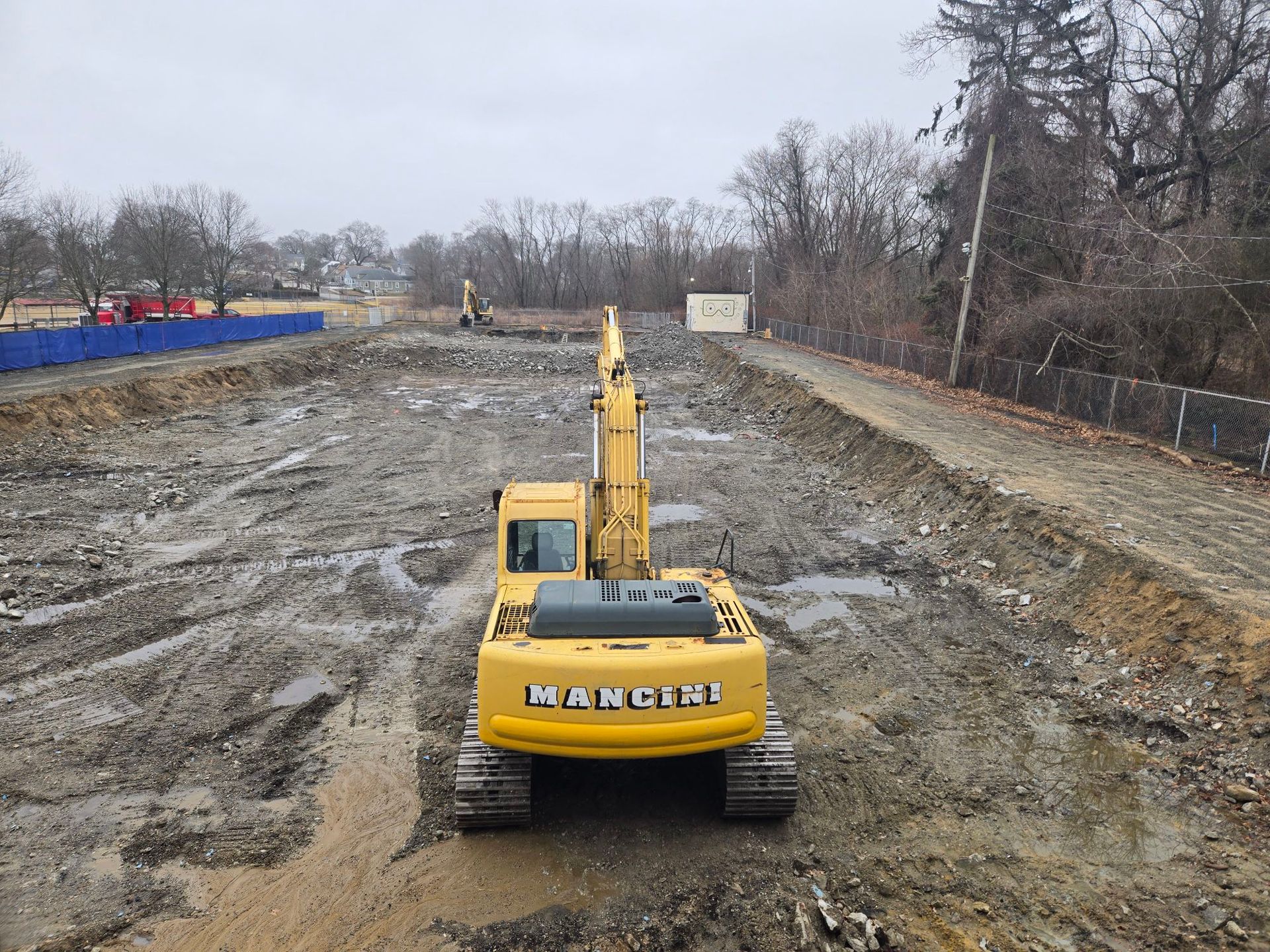 Yellow excavator on a muddy construction site with a dug-out trench and bare trees in the background