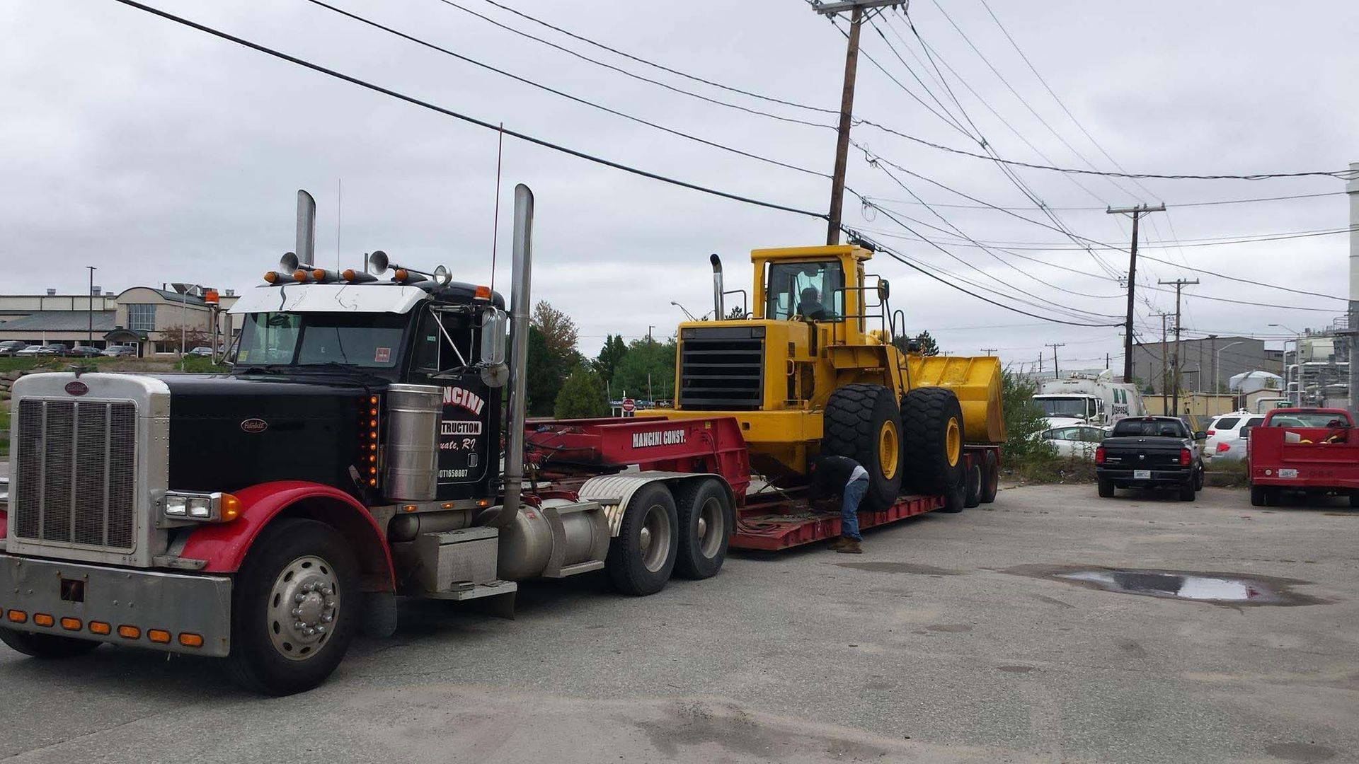 Red tow truck hauling a yellow construction vehicle on a city street under cloudy skies