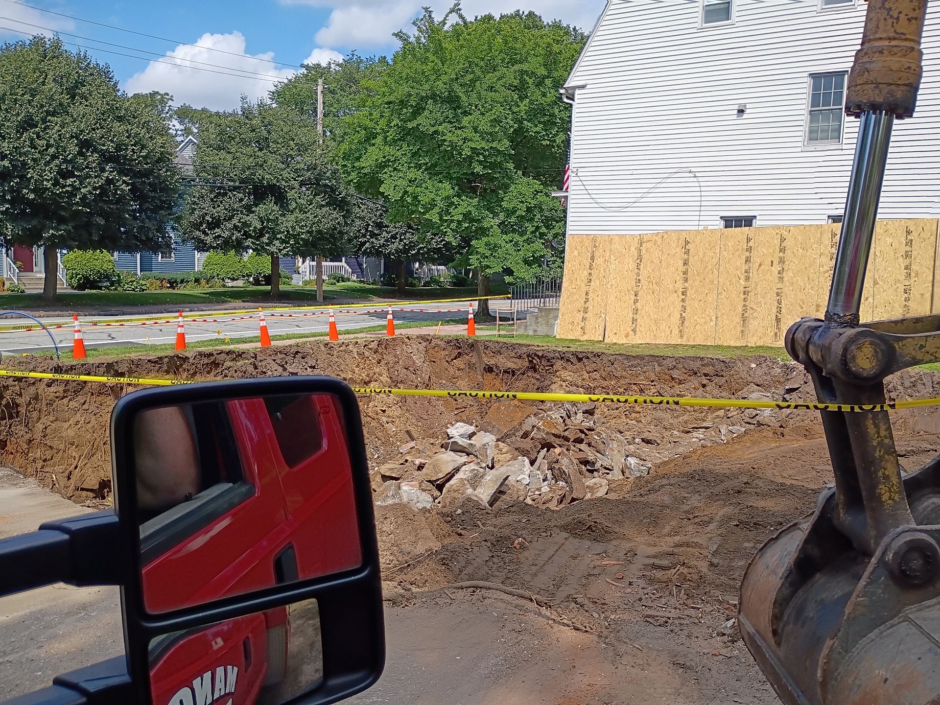Construction site with muddy ground, caution tape, orange cones, and a red vehicle mirror in the foreground
