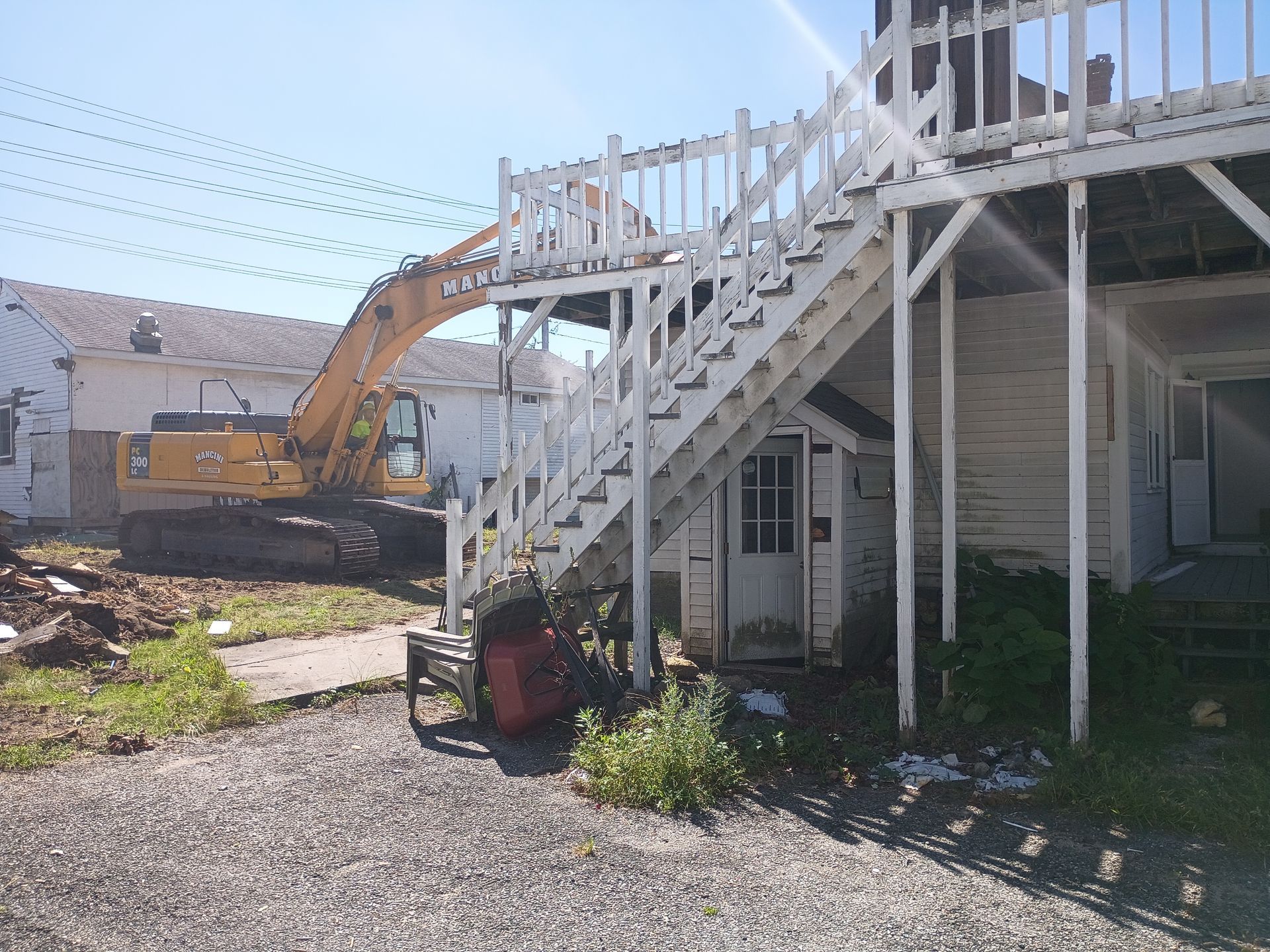 Excavator beside a white house with an exterior staircase, in a gravel yard under a clear sky
