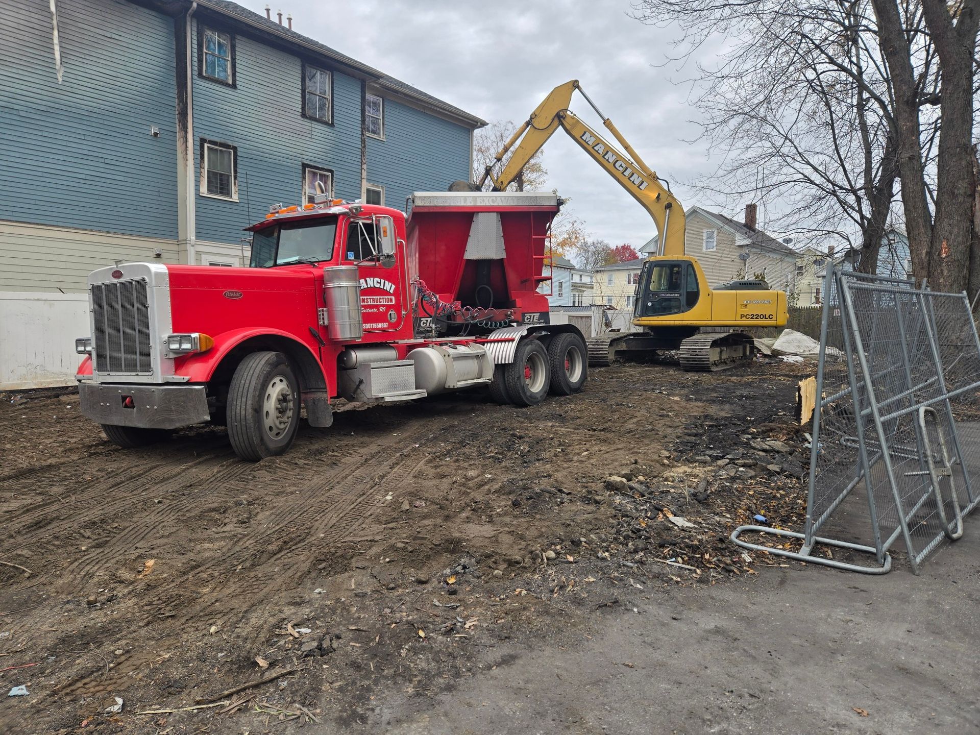 Red dump truck and yellow excavator on muddy construction site beside a blue building