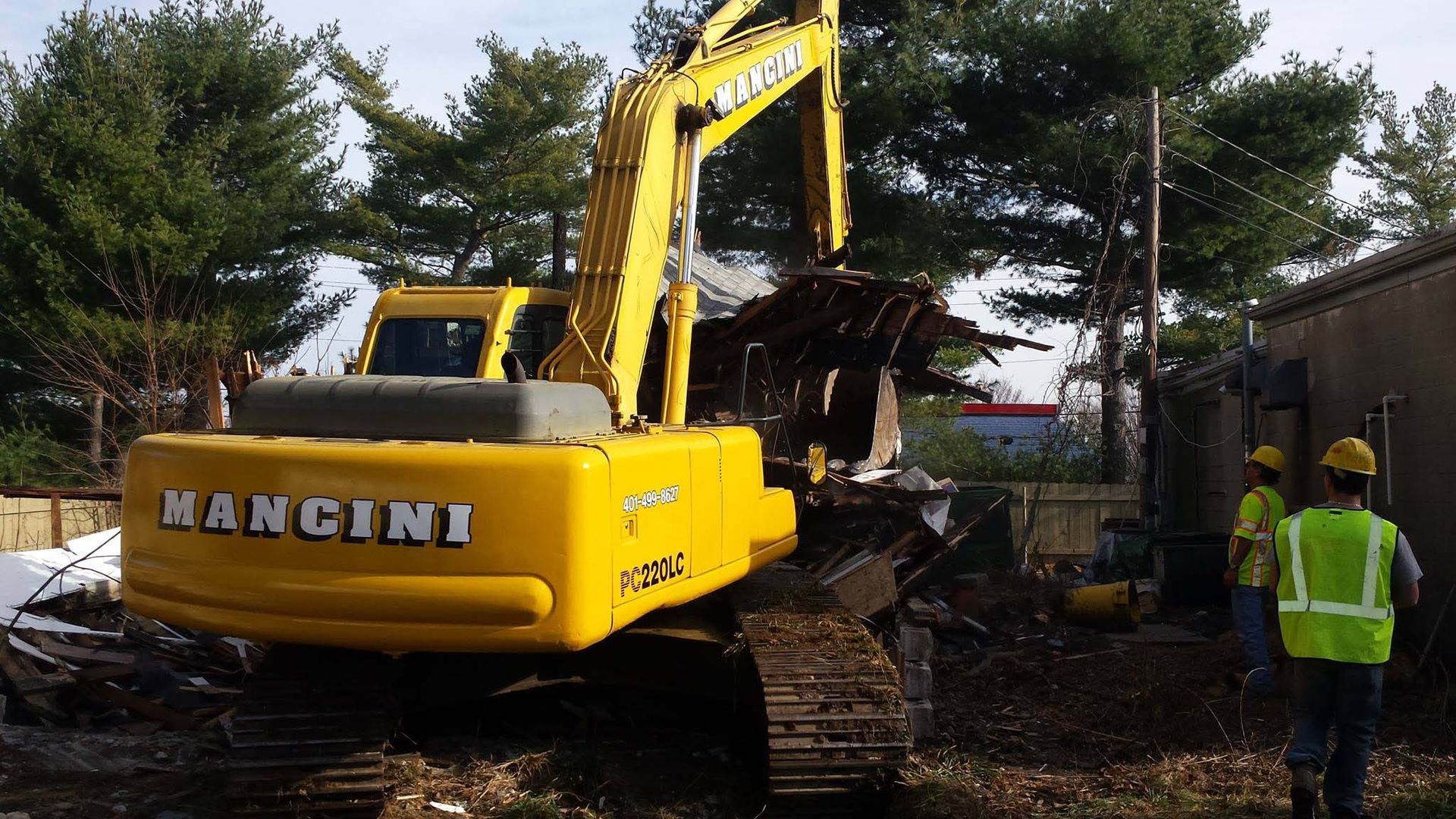 Yellow Mangini excavator lifting debris at a wooded demolition site, with a worker in a safety vest nearby