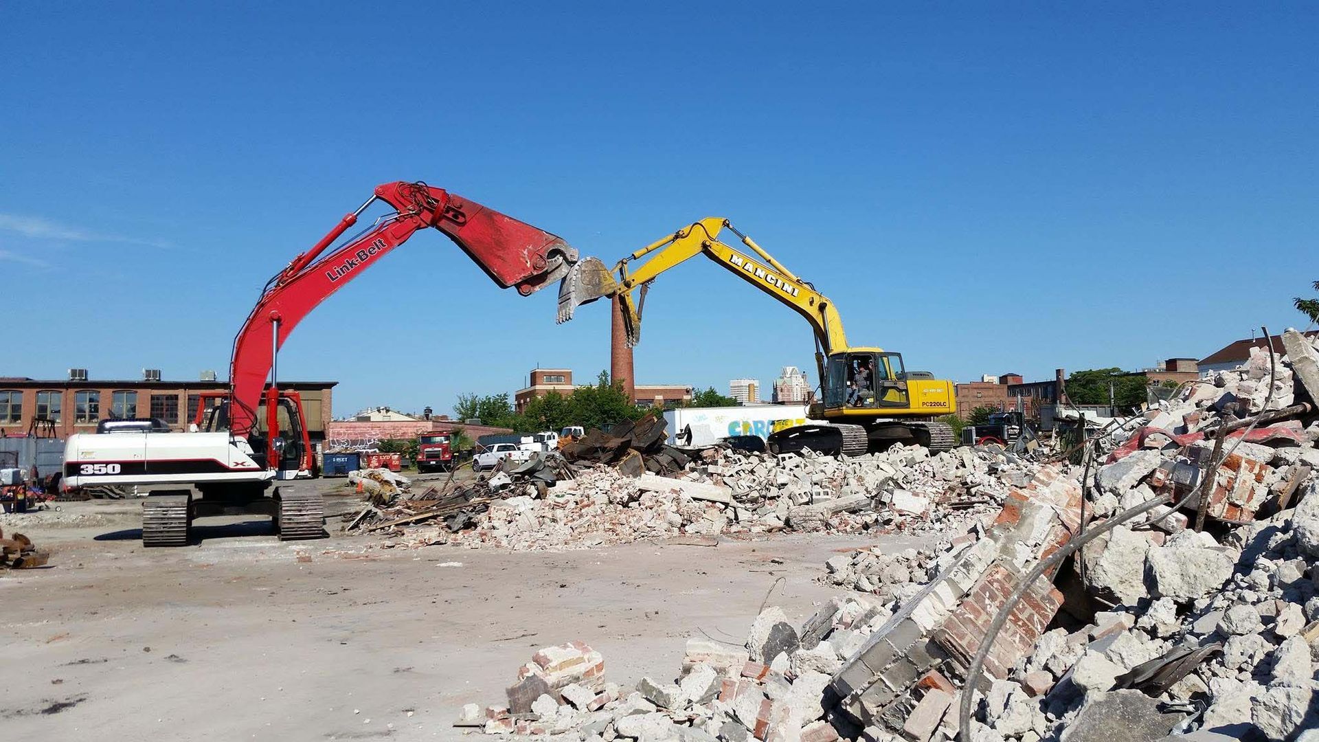 Two excavators clear rubble at a demolition site under a clear blue sky.