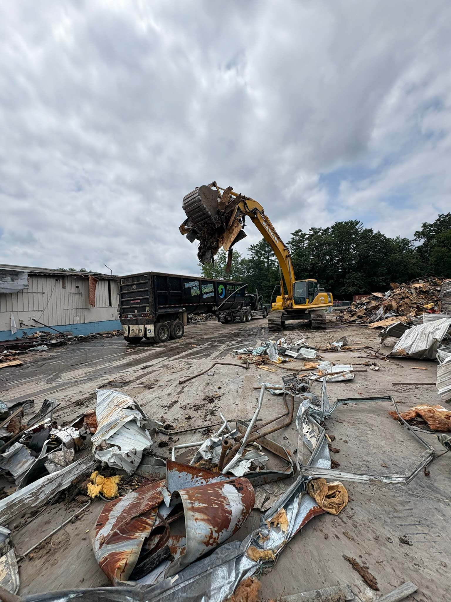 Excavator loading debris into a truck at a demolition site under a cloudy sky