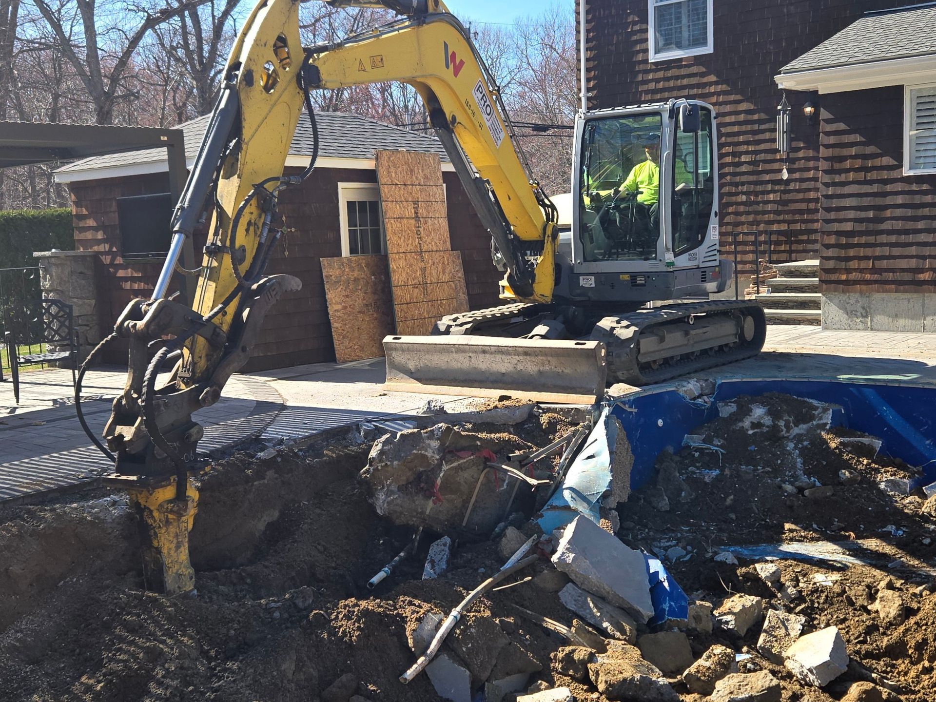 Yellow excavator demolishing debris beside a house on a street under bright sunlight