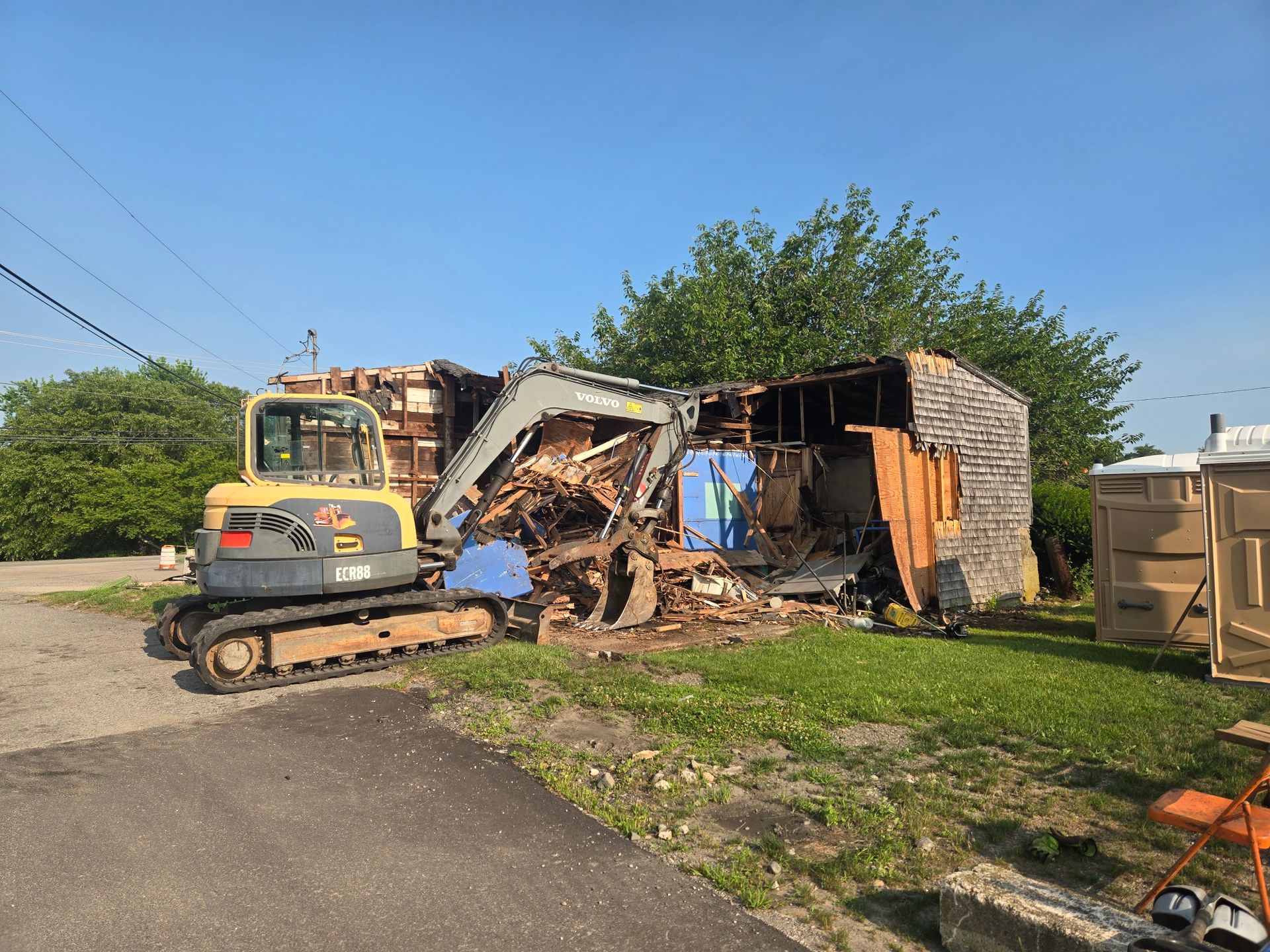 Excavator tears down a small shed beside a road, with piles of wood debris and a trailer nearby.