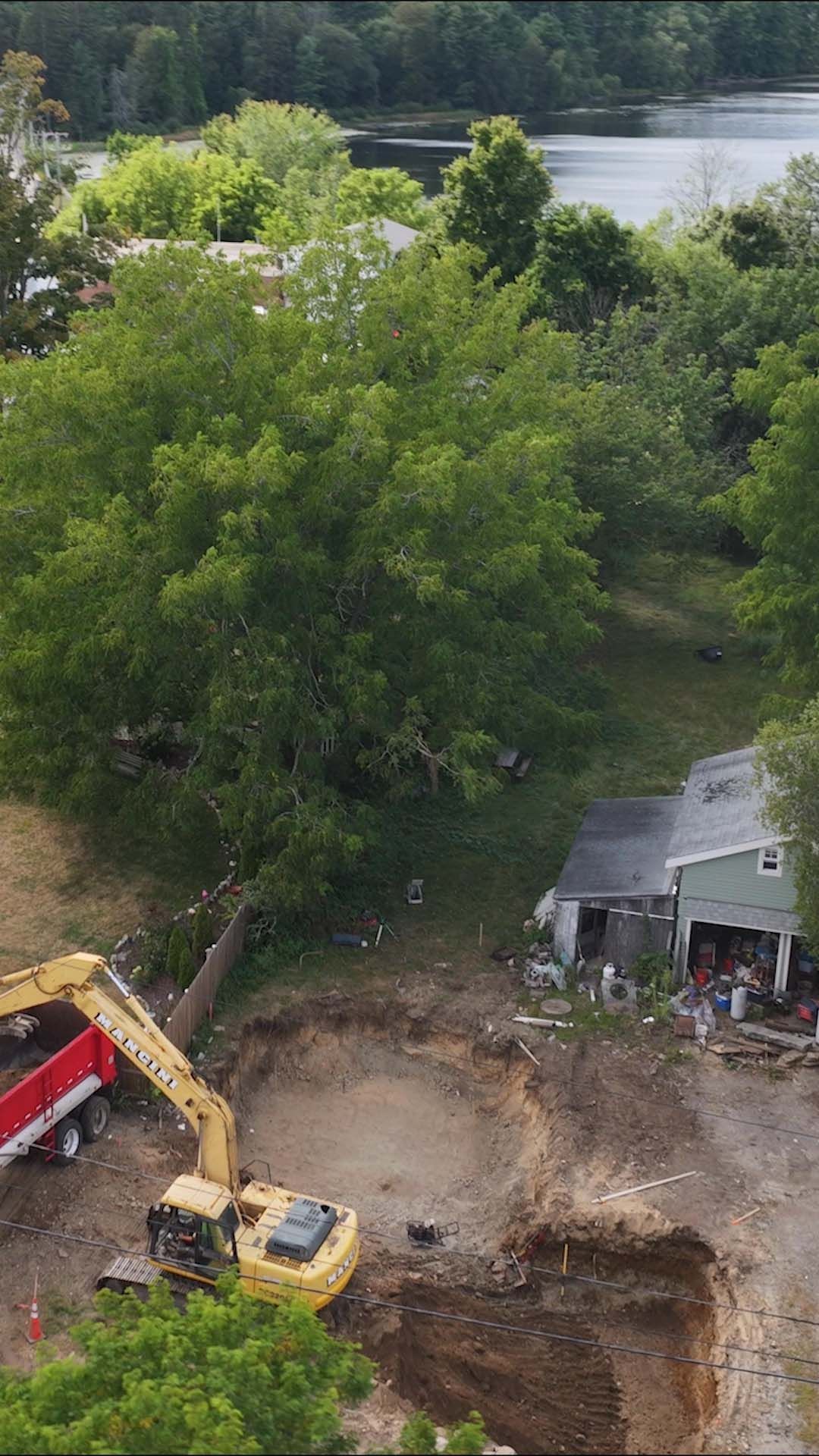 Excavator and dump truck beside a large dirt excavation near a house, with trees and water in the background