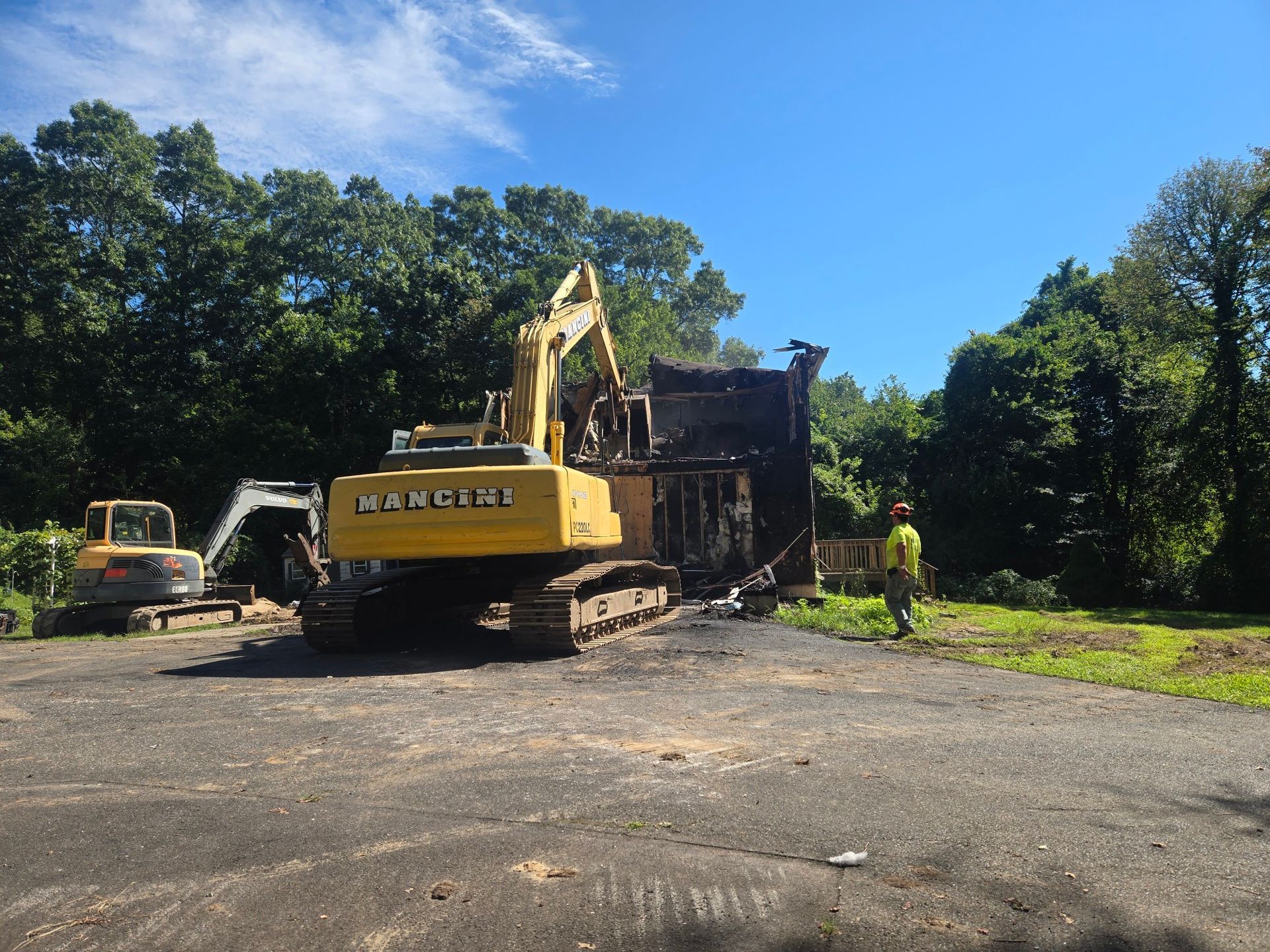 Yellow excavator at a wooded construction site, with workers clearing debris near a dark structure.