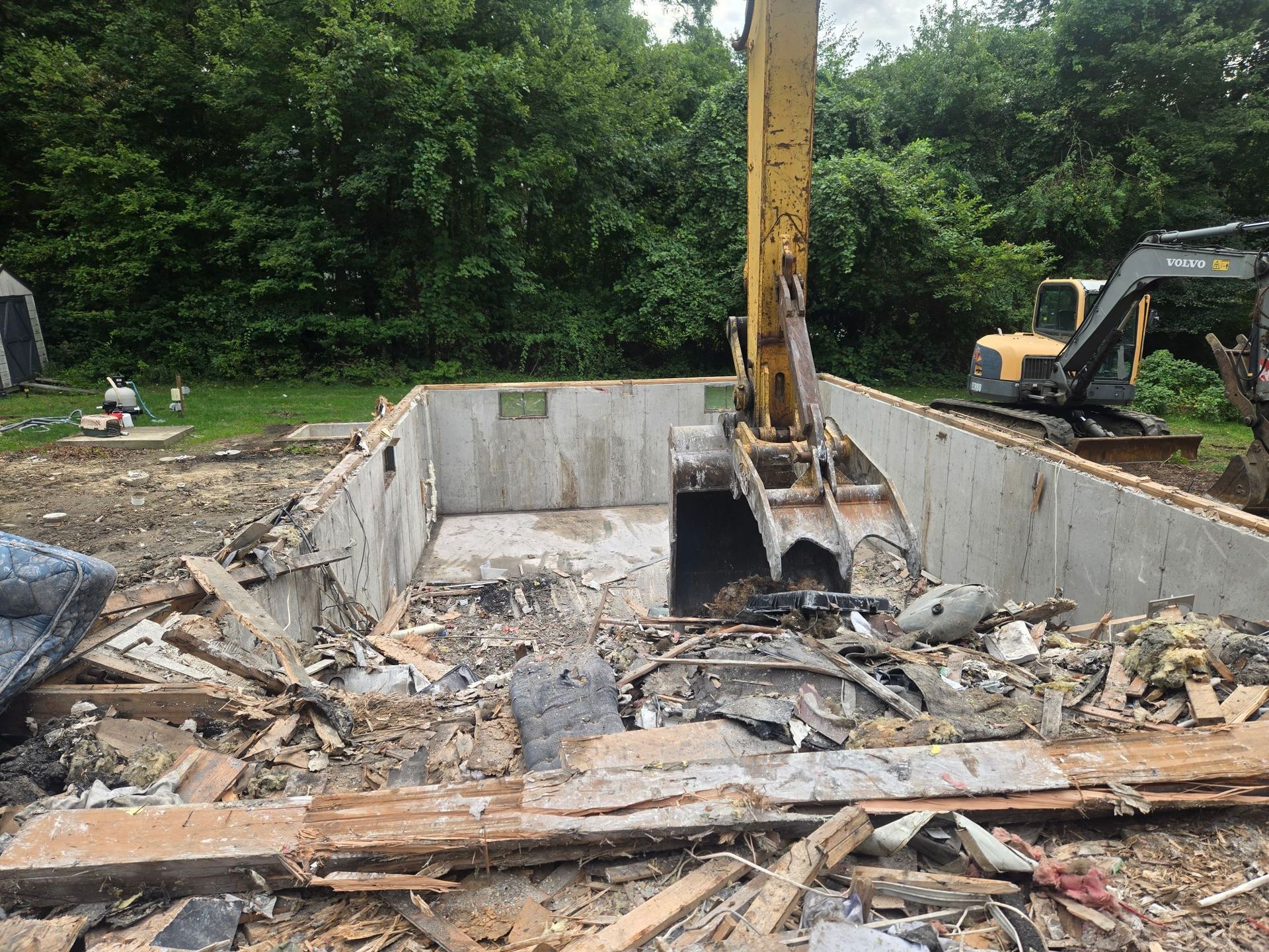 Excavator clearing debris in a concrete demolition pit with a second machine nearby and trees in the background